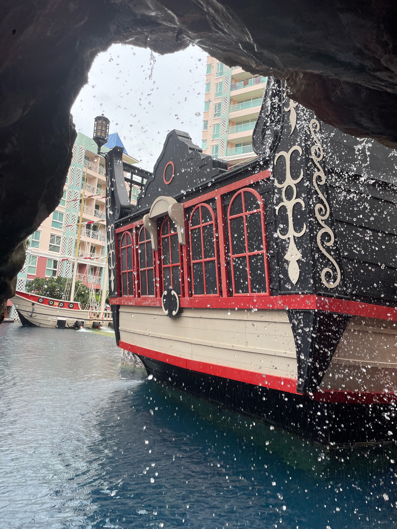 The image captures a view from within a rocky overhang, looking out onto a body of water. In the foreground, a portion of a black and red pirate-ship-themed structure is visible, with water droplets falling from the overhang onto its surface, creating splashes. In the background, a beige and red sailboat-style structure is docked, with several multi-story buildings featuring balconies and balconies visible behind it. The water appears calm with ripples. The overall scene suggests a themed attraction, possibly at a resort or water park, in Pattaya City, Thailand.