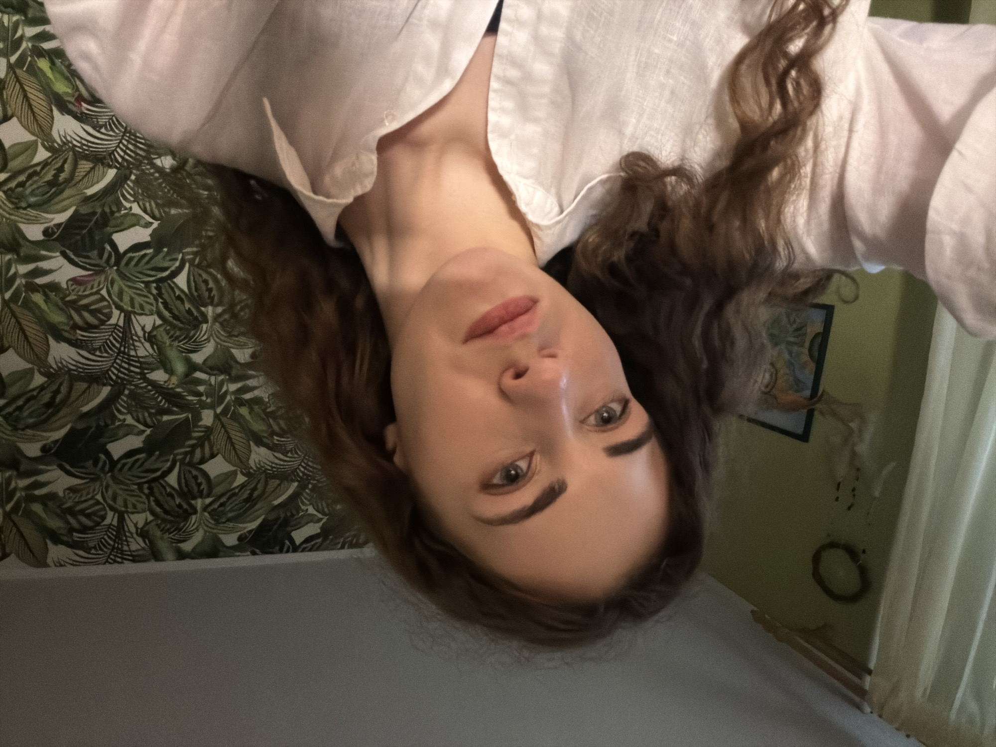 A young woman with long, wavy brown hair is looking upwards and slightly to the left, captured in a close-up, head-and-shoulders shot. She is wearing a white, collared shirt. The background features a wall with green paint on the left, adorned with a circular decorative item and a framed picture. To the right, the wall is covered with a patterned wallpaper depicting large green leaves, suggesting a tropical or botanical theme. The lighting appears to be soft, possibly natural light from a window. The scene is likely indoors. There is no discernible information to place this image in Zavodoukovsk, Russia.