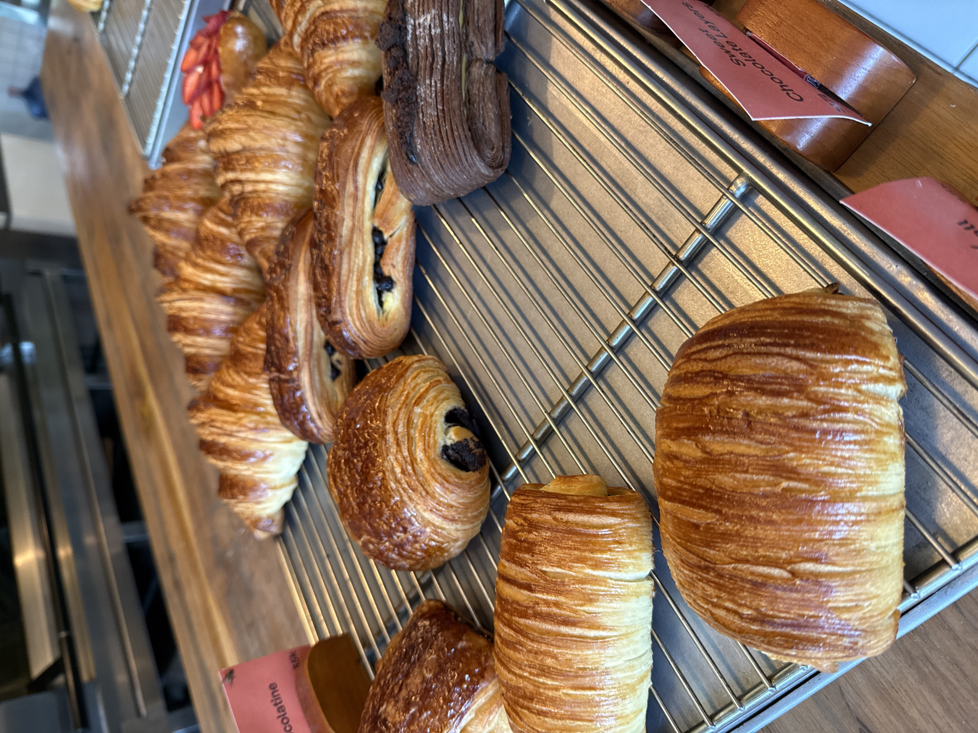 The image displays a close-up, eye-level view of a selection of freshly baked pastries arranged on a metal display rack, likely within a bakery or cafe in North Kuta, Indonesia. The pastries rest on a metal grate positioned above a warm-toned wooden counter or shelf.
The primary subjects are various types of golden-brown, flaky pastries, predominantly European-style. On the left, several crescent-shaped croissants are visible, showcasing their distinct layered textures and glossy finish. Further to the right and lower in the frame, multiple pain au chocolat are displayed, some with visible chocolate filling, alongside other cylindrical or spiraled layered pastries. A larger, beautifully browned and layered pastry, resembling a croissant or pain suisse, occupies a prominent position on the far right. A darker, chocolate-colored layered pastry is also visible in the upper-middle section.
A red, blurred object, possibly sliced fruit or a decorative element, is partially visible behind some croissants in the upper-left quadrant.
Small, pink paper tags are placed among the pastries, indicating their names or descriptions. Visible text includes "Sweet Chocolate Layers" on one tag and a partial word "...colatine" (likely "Chocolatine") on another, specifying the type of baked goods available. The pastries appear well-baked, with a rich, inviting color and texture, suggesting they are fresh for sale. The scene conveys an atmosphere of a well-stocked bakery display. No people are visible in the shot.
