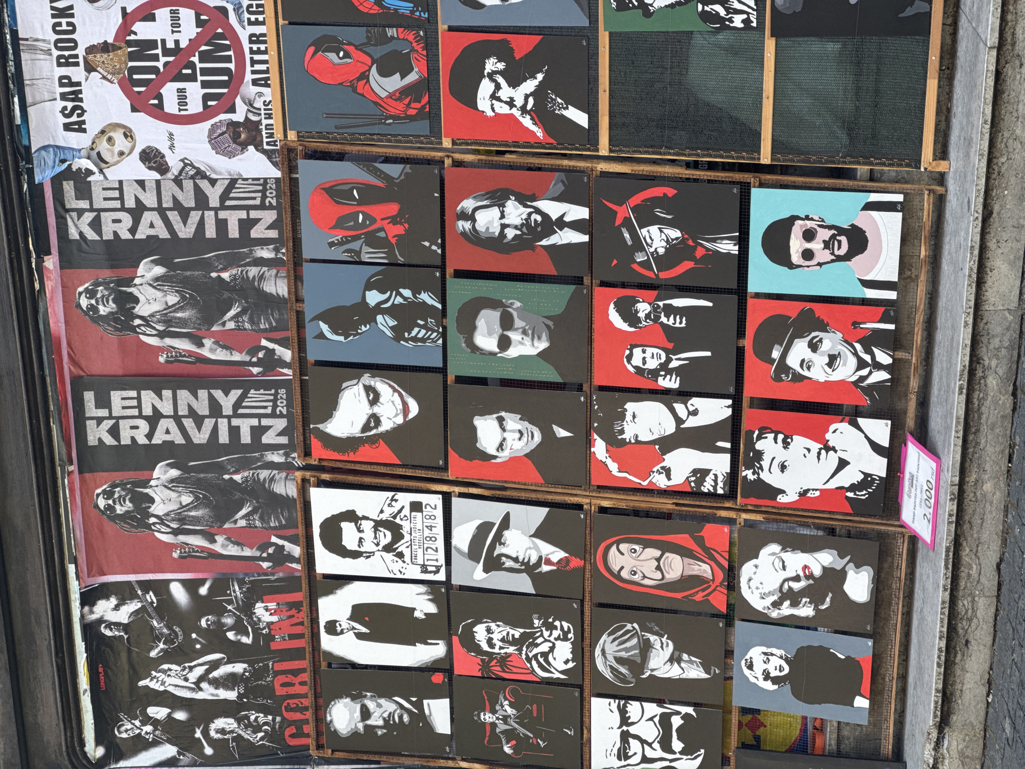 The image captures a vibrant street art display in Belgrade, Serbia, likely located near a tourist-heavy area. The foreground features a row of colorful paintings, predominantly pop art portraits, displayed on wooden racks set up on a stone sidewalk. To the right, a pink sign indicates "HAND PAINTED POP-ART PAINTING" and lists a price of "2.000 rsd."
The paintings showcase a diverse range of iconic figures from popular culture and film. Among them are portraits of actors like Al Pacino, Robert De Niro, and Keanu Reeves, as well as characters such as Batman, Deadpool, and the Joker. Other notable subjects include Marilyn Monroe, Audrey Hepburn, Charlie Chaplin, and the character of Dali from "Money Heist." The art style is characterized by bold lines, flat colors, and simplified features, creating a striking visual impact.
Behind the paintings, a series of large banners hang, advertising concerts and events. One prominent banner features a black and white image of Lenny Kravitz with the text "LENNY KRAVITZ LIVE 2026." Another banner displays a band performing, possibly "Goblin." Further to the right, a banner advertises "A$AP ROCKY" and features a graphic with a "no tour" symbol. The overall scene suggests a bustling marketplace or an area dedicated to selling art and memorabilia to passersby. The lighting indicates it is daytime, with natural light illuminating the display.