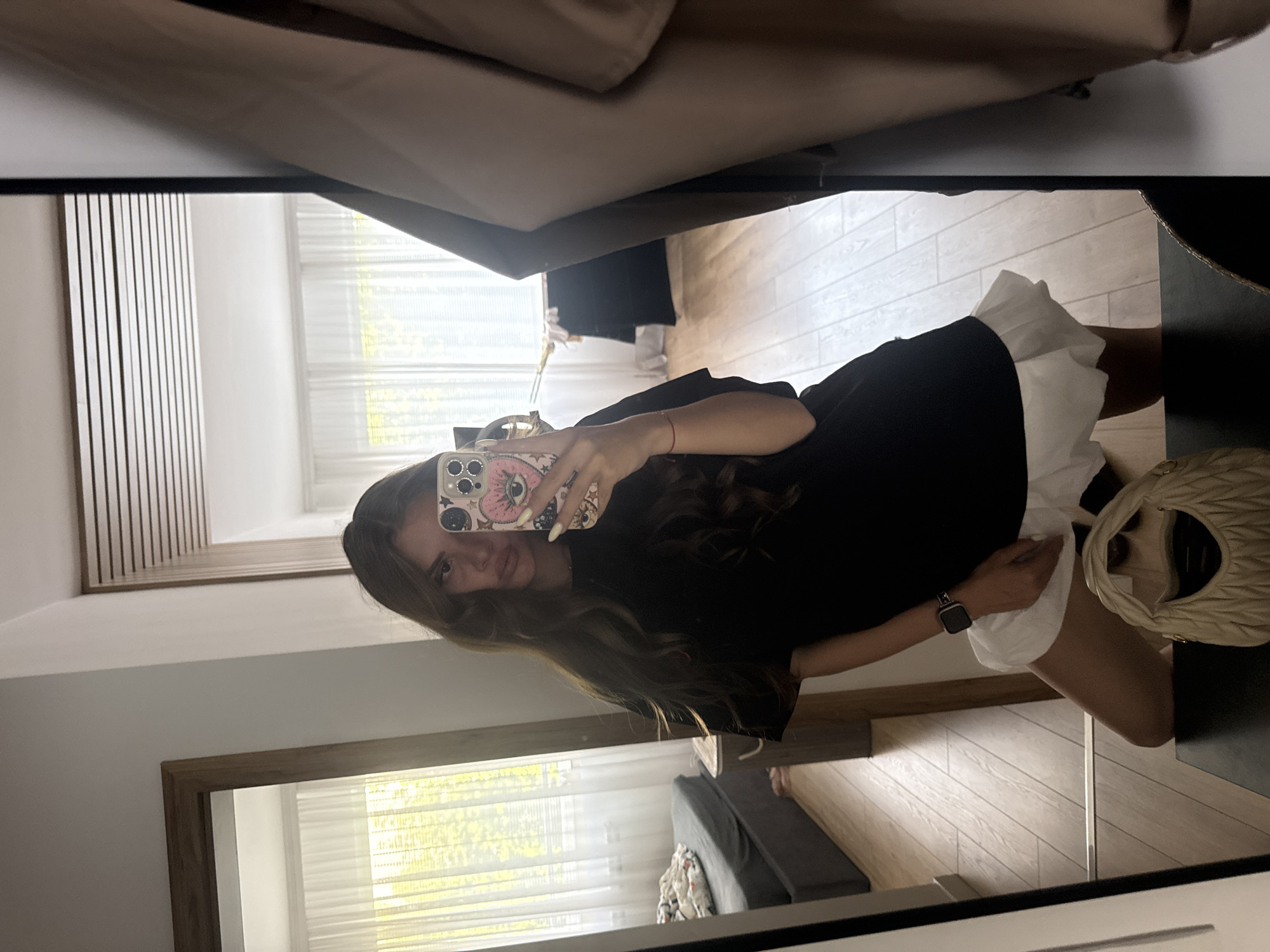A young woman with long brown hair takes a mirror selfie in what appears to be a bedroom or dressing area. She is wearing a black dress with a white ruffled hem and a smartwatch on her left wrist. She holds a smartphone with a decorative case in her right hand. A light-colored, textured handbag is visible next to her on a dark surface.
The background shows a wooden floor and white blinds covering a window, suggesting an indoor setting. In the reflection of another mirror, a sliver of a couch or daybed is visible, along with a framed artwork or headboard. The overall lighting suggests it's daytime. The woman's expression is neutral as she poses for the photo.