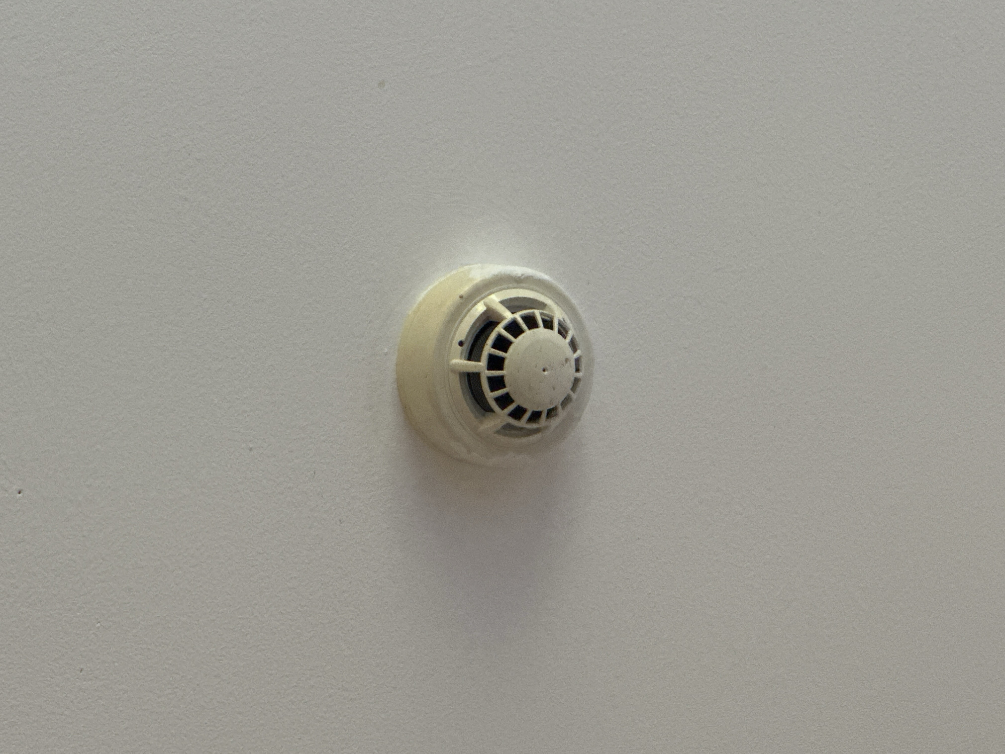 A smoke detector is mounted on a white textured ceiling. The smoke detector is round and cream-colored with a patterned grille on its surface. It is positioned centrally in the frame, with a slight shadow cast to its left. The ceiling appears to be part of an indoor environment.