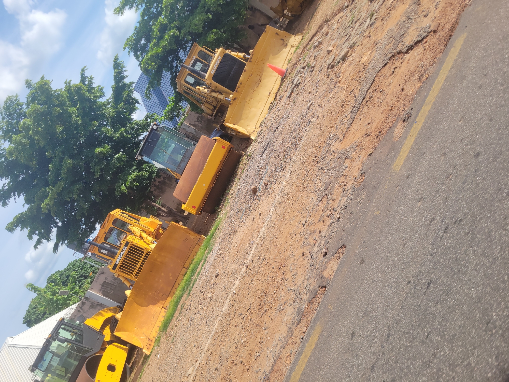The image shows a construction site in Lokoja, Nigeria. Several yellow construction vehicles, including bulldozers and a roller, are parked on a dirt and gravel surface adjacent to a paved road. Trees and a modern building with blue, glass-like panels are visible in the background. The sky is partly cloudy, suggesting it is daytime. The overall scene depicts an active or recently used construction area. There is no visible text in the image.