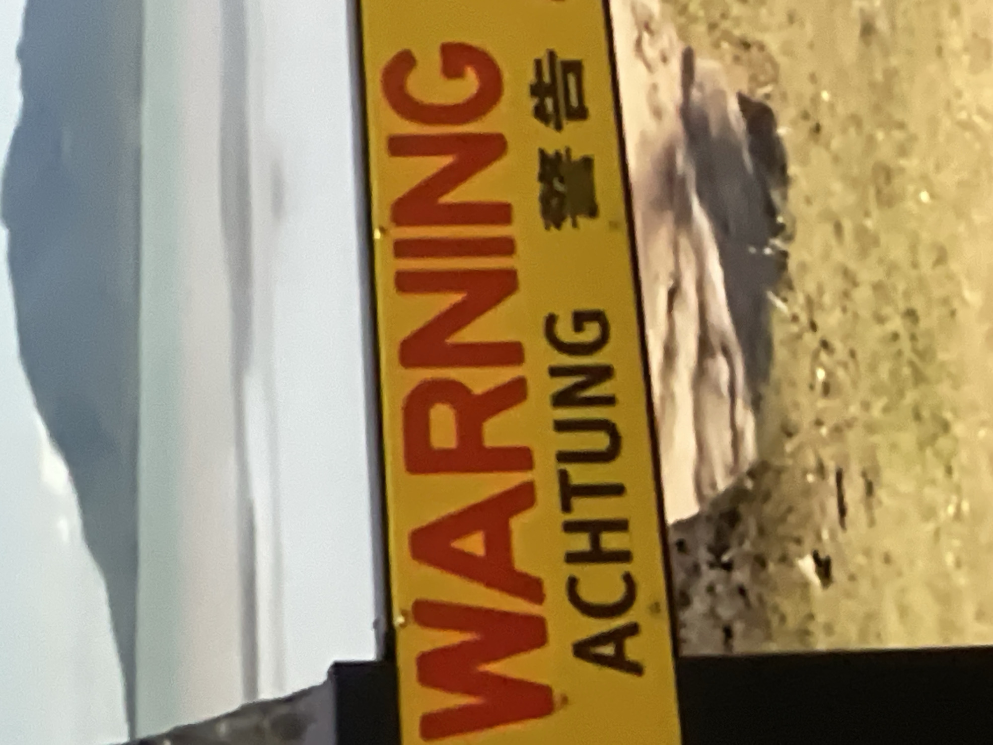 A yellow warning sign with red lettering is affixed to a black pole. The sign reads "WARNING" in large letters across the top. Below that, in smaller letters, it says "ACHTUNG" followed by what appears to be Japanese or Chinese characters. The background of the sign is a blurry image of water and a distant mountain range. The foreground shows grassy ground and rocks. The image does not provide any information that would allow for identification of the location as Pattaya City, Thailand, or any specific city.