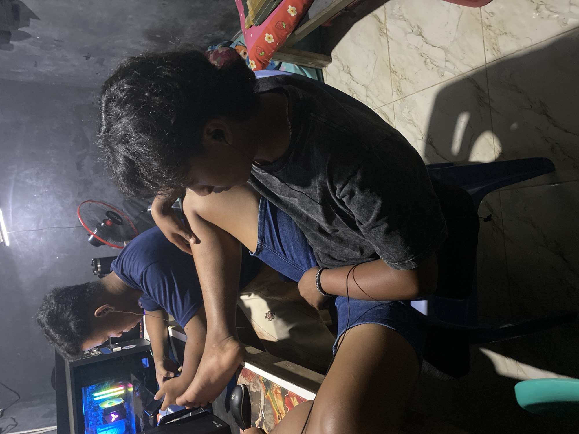 In the dimly lit interior of what appears to be a residential room in Sumber Baru, Indonesia, two young individuals are engaged with a computer setup. The room's walls are dark and textured, and overhead lighting casts long shadows on the patterned floor.
In the foreground, a person with dark, curly hair is seated, facing away from the camera and slightly to the left, their attention focused on a smartphone held in their hands. They are wearing a dark grey t-shirt and denim shorts, with a beaded bracelet on their left wrist. A thin black wire, likely from earbuds, is visible extending from their ear.
Behind and to the left of the foreground individual, another person is seated, also facing the computer screen. They are wearing a blue t-shirt and are holding a smartphone. Their left foot is resting on the edge of a desk. The computer monitor displays a colorful, illuminated scene, possibly a game. A black computer mouse is visible on the desk. A black oscillating fan with a red trim is positioned in the background, to the right of the computer setup, indicating a warm climate. The overall impression is of a casual, indoor activity, likely gaming or browsing on electronic devices.