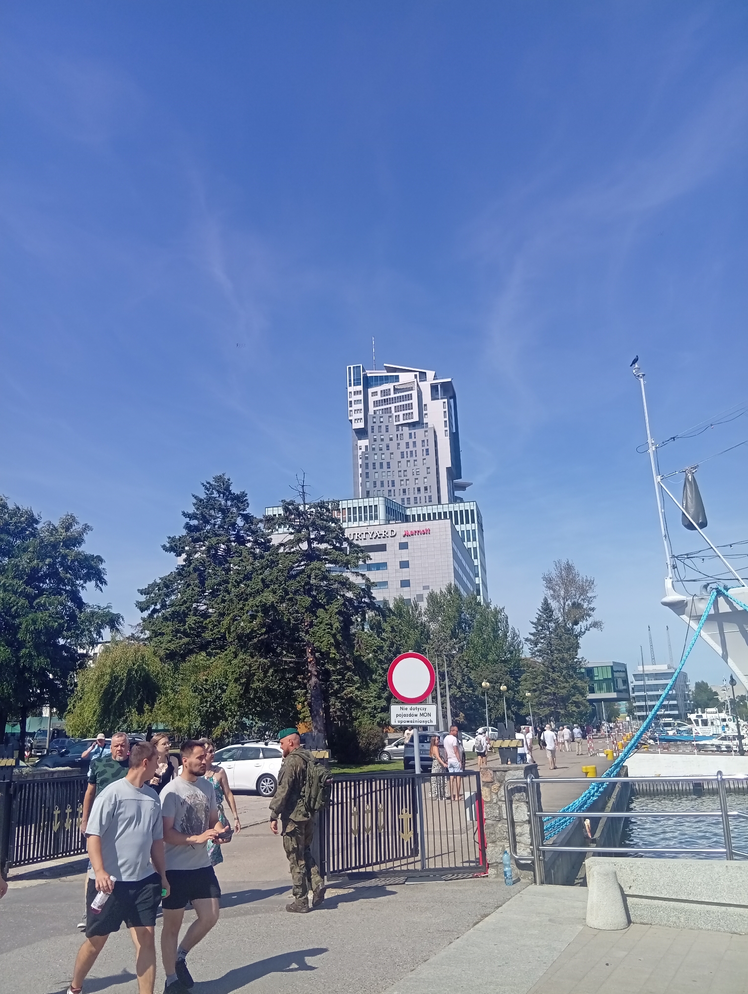 In Gdynia, Poland, a clear blue sky with wispy clouds forms the backdrop for a scenic urban view. In the distance, the distinctive multi-story facade of the Sea Towers complex, a prominent landmark in the city, rises majestically. The lower section of the complex displays the logos "COURTYARD Marriott".
In the foreground, a group of people strolls along a paved walkway adjacent to a marina. The walkway is bordered by a black metal fence with anchor motifs. A uniformed soldier in camouflage stands guard near the entrance to the walkway. To the right, a large ship, with a blue rope prominently displayed, is moored in the water. Lush green trees provide a natural contrast to the urban architecture. A circular red and white sign, a traffic regulatory symbol, is visible on the left side of the walkway. Several cars, including a white SUV and other vehicles, are parked along the street in the background. The sun casts long shadows, indicating a bright, daytime scene.