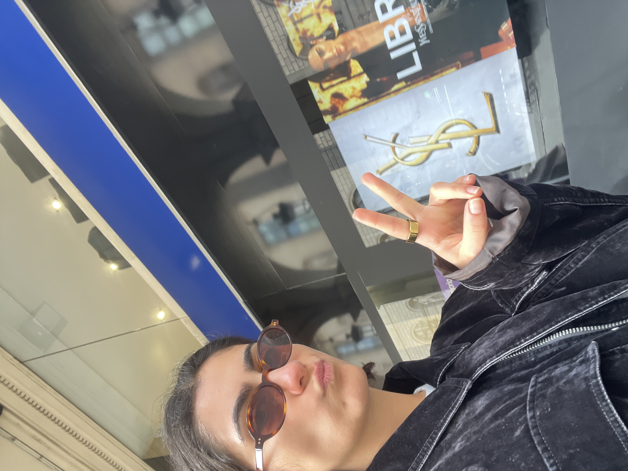 A young adult, appearing to be female, is depicted in a selfie, looking directly at the camera with a playful expression, her lips slightly pursed in a pout. She wears round, tortoiseshell-patterned sunglasses and a dark, possibly velvet or corduroy, jacket. Her right hand is raised, making a peace sign, and she sports a gold-colored ring on her ring finger.
The background is a reflective surface, possibly a window or a large digital screen. Reflected elements include abstract shapes, bright round lights, and what appears to be the interior or exterior of buildings. A distinct blue stripe is visible on the left side of the reflection.
Behind the person's hand, an advertisement is prominently displayed on the reflective surface. It features the bold text "LIBRE" at the top, along with the iconic gold "YSL" (Yves Saint Laurent) logo. Part of a person's image is also visible within the advertisement. The setting, with the luxury fashion advertisement, suggests a commercial area, possibly a high-end shopping district or department store. The lighting indicates it is likely daytime, either indoors or in a covered outdoor space.