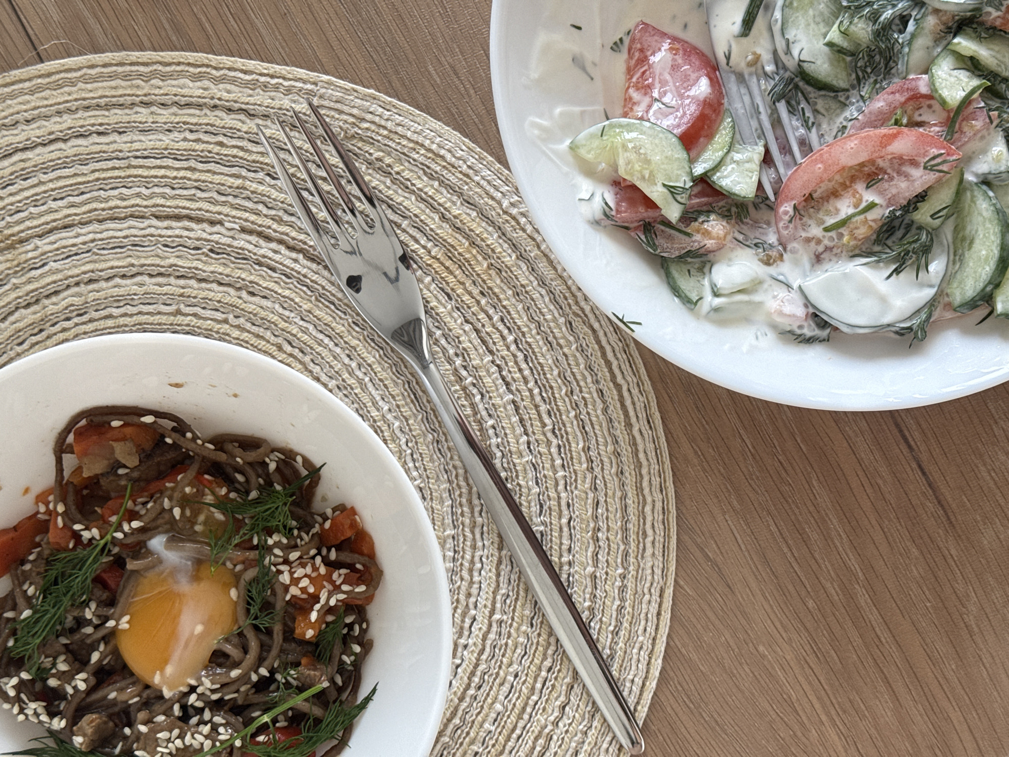 The image shows a wooden table setting with a round woven placemat. On the left side of the placemat is a white bowl containing soba noodles mixed with vegetables, sesame seeds, and topped with an egg yolk and dill. A silver fork lies diagonally across the placemat. To the upper right is a white bowl with a salad of sliced tomatoes and cucumbers in a creamy dressing with dill. The location is unknown.