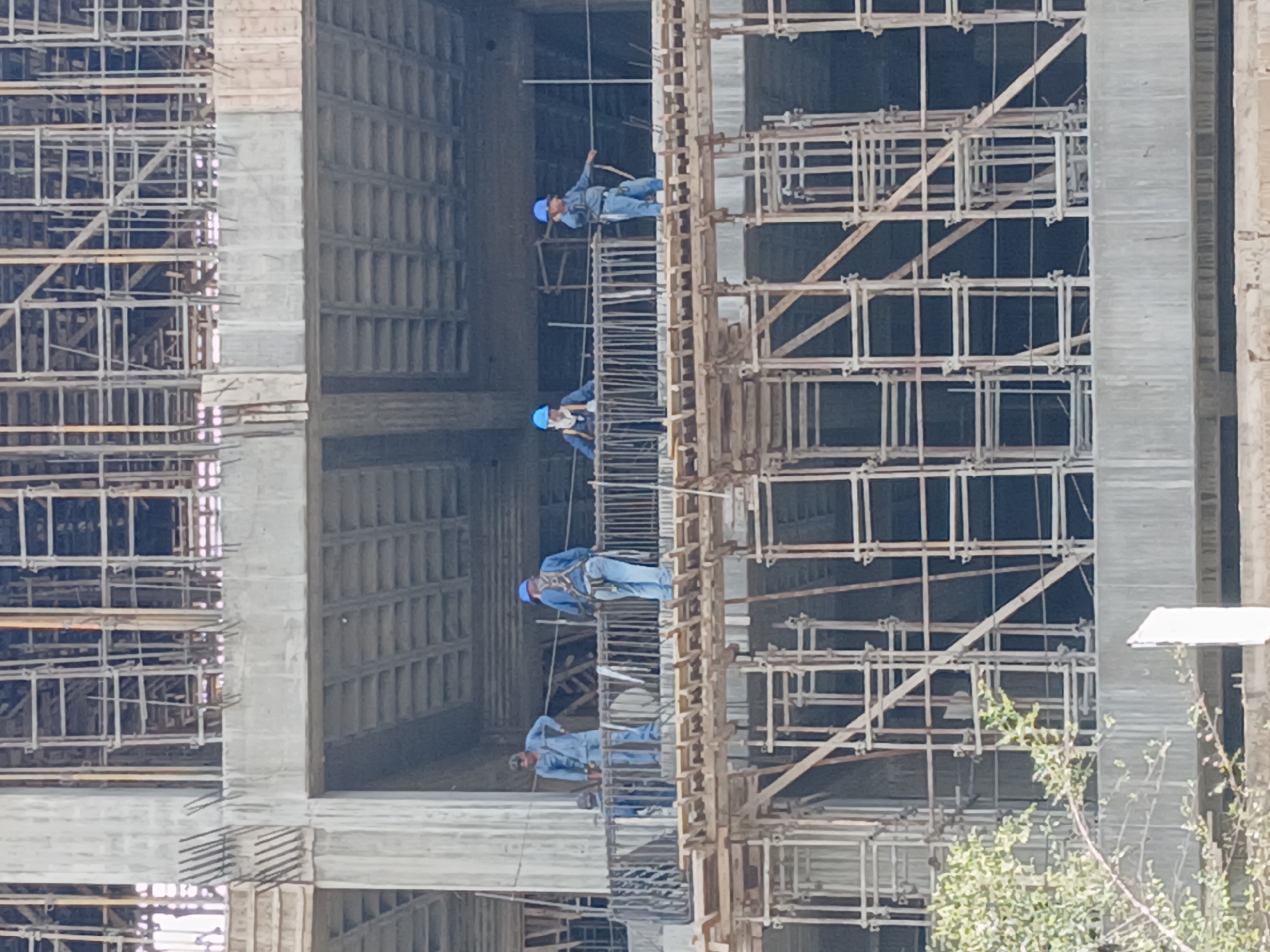 The image shows a construction site, likely a high-rise building under construction, featuring scaffolding and construction workers. The workers are wearing blue helmets and work clothes, and are positioned on the scaffolding at different levels. The building is primarily made of gray concrete and steel. The location is indicated as Ciudad Guayana, Venezuela, based on the prompt. The sky is light gray, suggesting overcast conditions. Some green foliage is visible at the bottom right, indicating vegetation in the surrounding area.