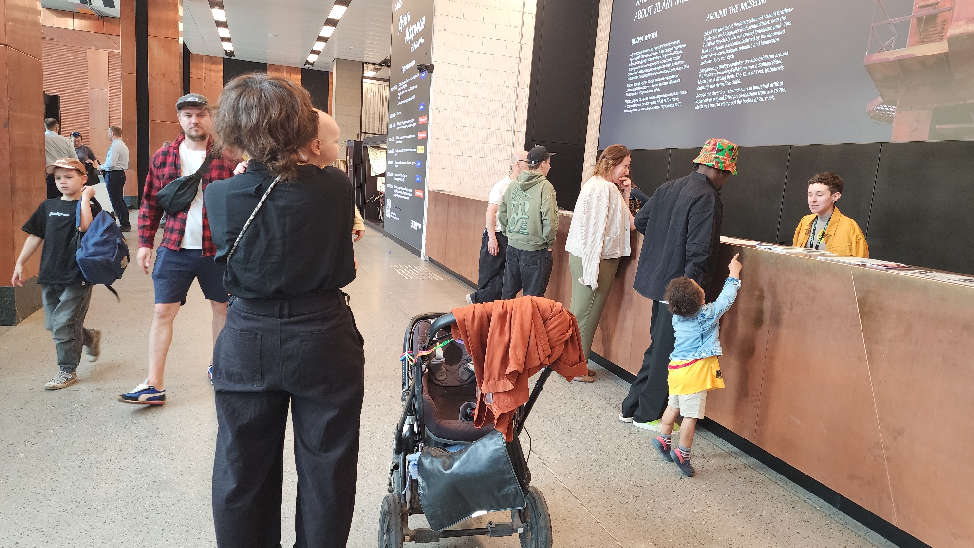 The image shows the interior of ZILART, a museum located at the intersection of Vesnin Brothers Boulevard and Alexander Rodchenko Street, Moscow, Russia. Several people are present in the scene. In the foreground, a woman with dark clothing holds a baby, standing near a stroller. To their left, a young boy with a backpack walks alongside a man in a red flannel shirt. To their right, several adults are near the reception desk, where a museum employee stands ready to assist visitors. A young child points to the counter while interacting with the employee. Behind the desk is a board with text and illustrations describing the museum. The floor is light-colored, and the lighting is bright.