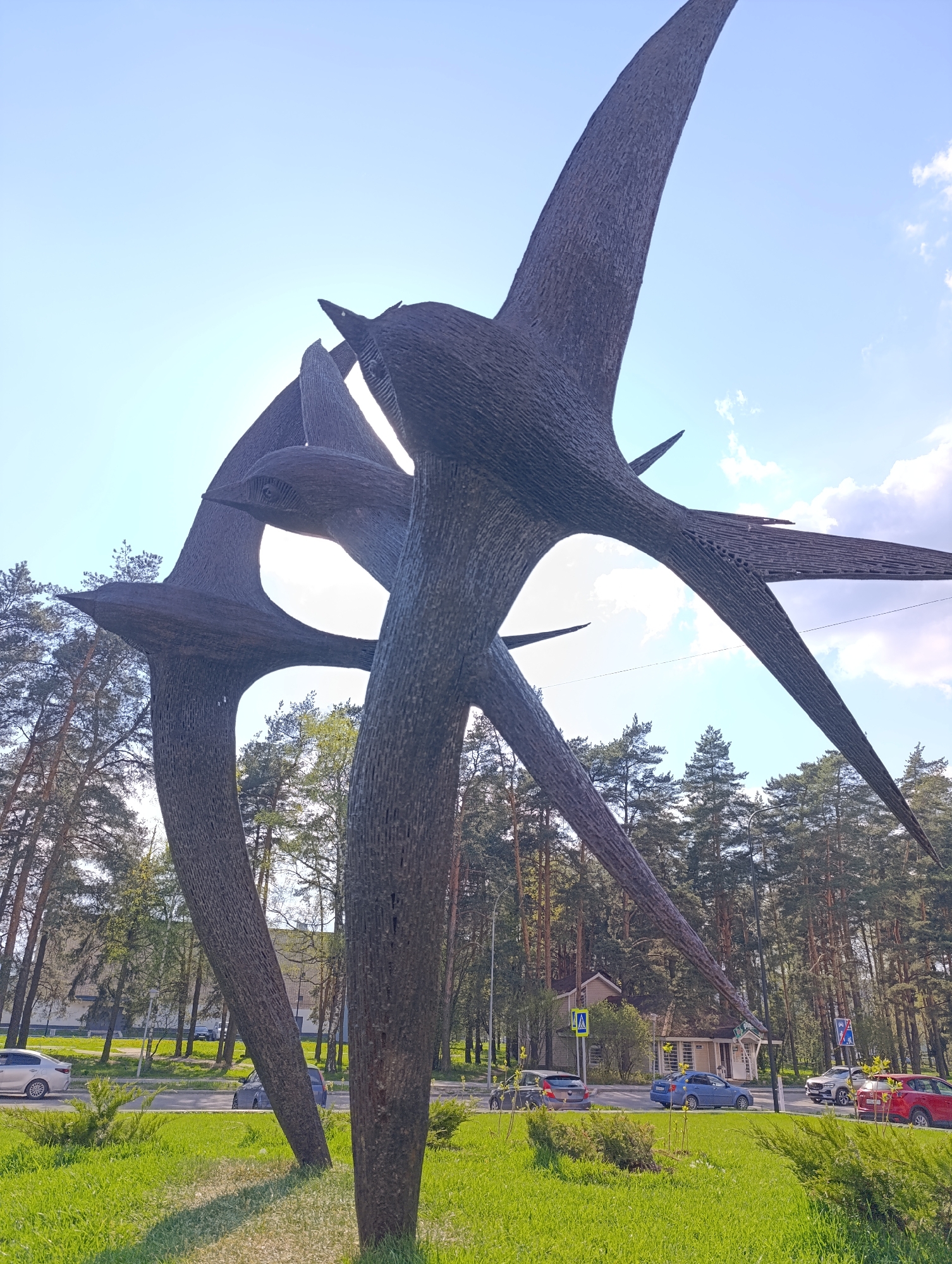 The image shows a large, dark-colored metal sculpture depicting birds in flight, situated on a grassy area. The location appears to be a park or green space within a city. Trees are visible in the background, and a street with cars parked along the roadside is present. In the background, there is a building with a crosswalk sign in front. The sky is blue with some clouds.