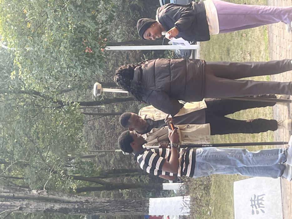 Four people stand in a park-like setting with trees in the background. One person wears a black beanie, black jacket, and lilac sweatpants, looking at a paper. Another individual has long, curly hair and wears a black puffer jacket and dark pants. Two men are closer, one in a striped shirt and jeans, and the other in a tan jacket and dark pants. The men appear to be looking at a phone. A white sign with a black symbol stands in the foreground.
