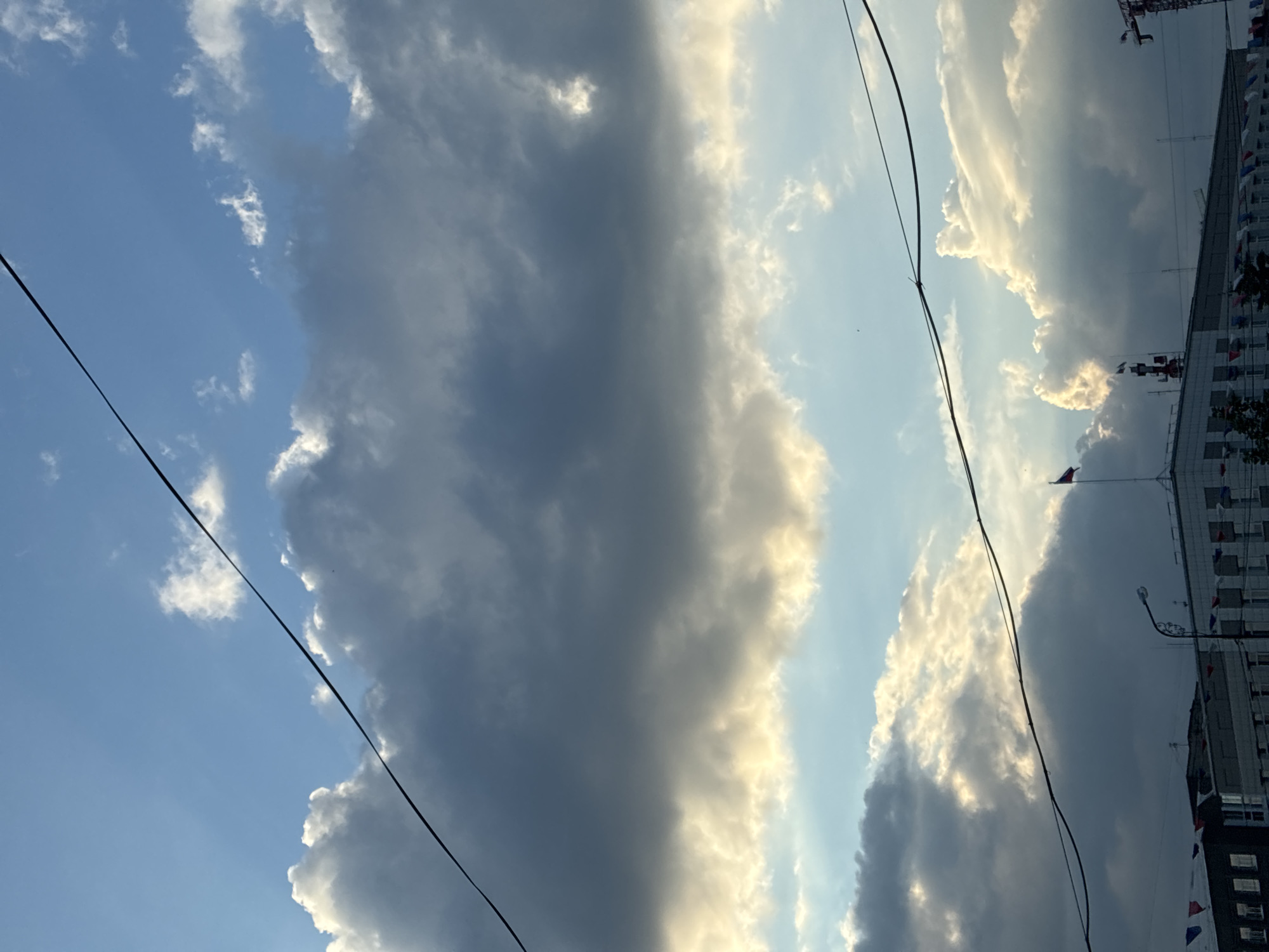 The image captures a dramatic sky over a city, likely Irkutsk, Russia, with a mix of dark, brooding storm clouds and bright, illuminated cumulus clouds. The sun's rays are breaking through, casting a golden glow on the edges of the clouds and illuminating patches of the blue sky.
In the foreground, the silhouettes of buildings are visible, along with various structures on their rooftops, including antennas and a flag. One flag, appearing to be the Russian flag, is prominently displayed on a flagpole. Wires stretch across the frame, adding a sense of urban infrastructure to the scene. Decorative flags or pennants, possibly of red, white, and blue, are strung along the roofs of some buildings, suggesting a festive or official occasion. A construction crane is visible on the far right. The overall impression is one of dynamic weather conditions over a cityscape.