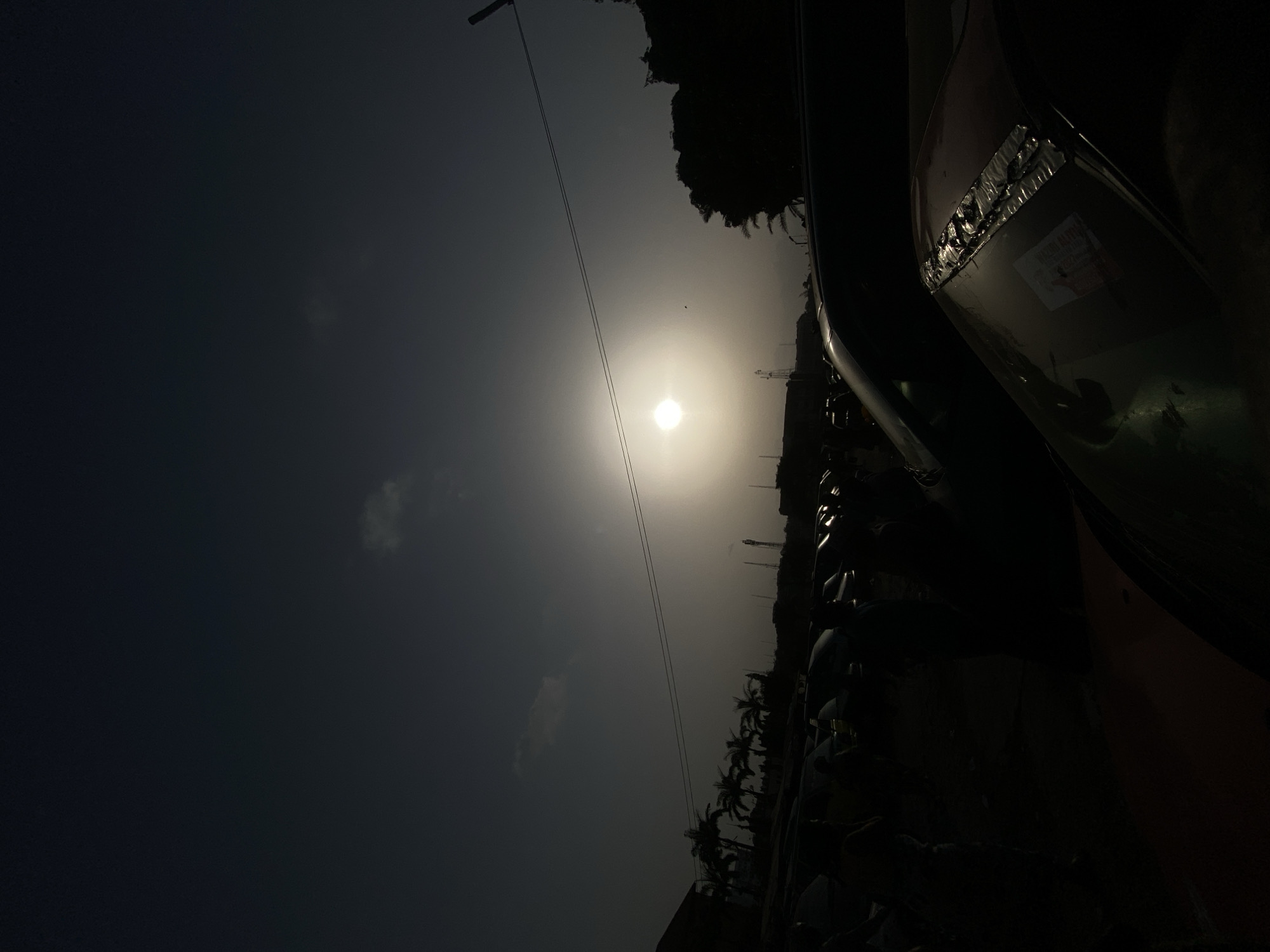 The image, oriented correctly, captures an outdoor scene at what appears to be midday or early afternoon in Maiburiji, Nigeria. A brilliant sun dominates the upper center of the frame, radiating intense light and creating a significant glare. The sky transitions from a dark, hazy grey in the upper left to a brighter, almost white hue around the sun, with a few faint clouds visible in the darker regions. Two thin, dark lines, resembling power lines, stretch diagonally across the sky from the upper left towards the sun, possibly connected to a pole that is mostly outside the frame. A small dark speck, likely a bird, is discernible near the sun.
The lower half and right side of the image are rendered in deep silhouette due to the strong backlighting. A distinct horizon line runs across the lower right, featuring a varied landscape of silhouetted trees, including several palm trees, interspersed with indistinct structures and poles. In the immediate foreground on the far right, a large, dark, angled object, possibly the side or bumper of a vehicle such as a truck or bus, is prominent. This object exhibits a section of crinkled, shiny, metallic-looking tape or material. A light-colored sticker is affixed to the vehicle, partially visible despite the heavy shadow. Text on this sticker includes "YAYO ALIYU" and a partial sequence that appears to be a phone number or address, starting with "0802". The overall impression is a high-contrast, atmospheric view of a bright day with strong directional light.