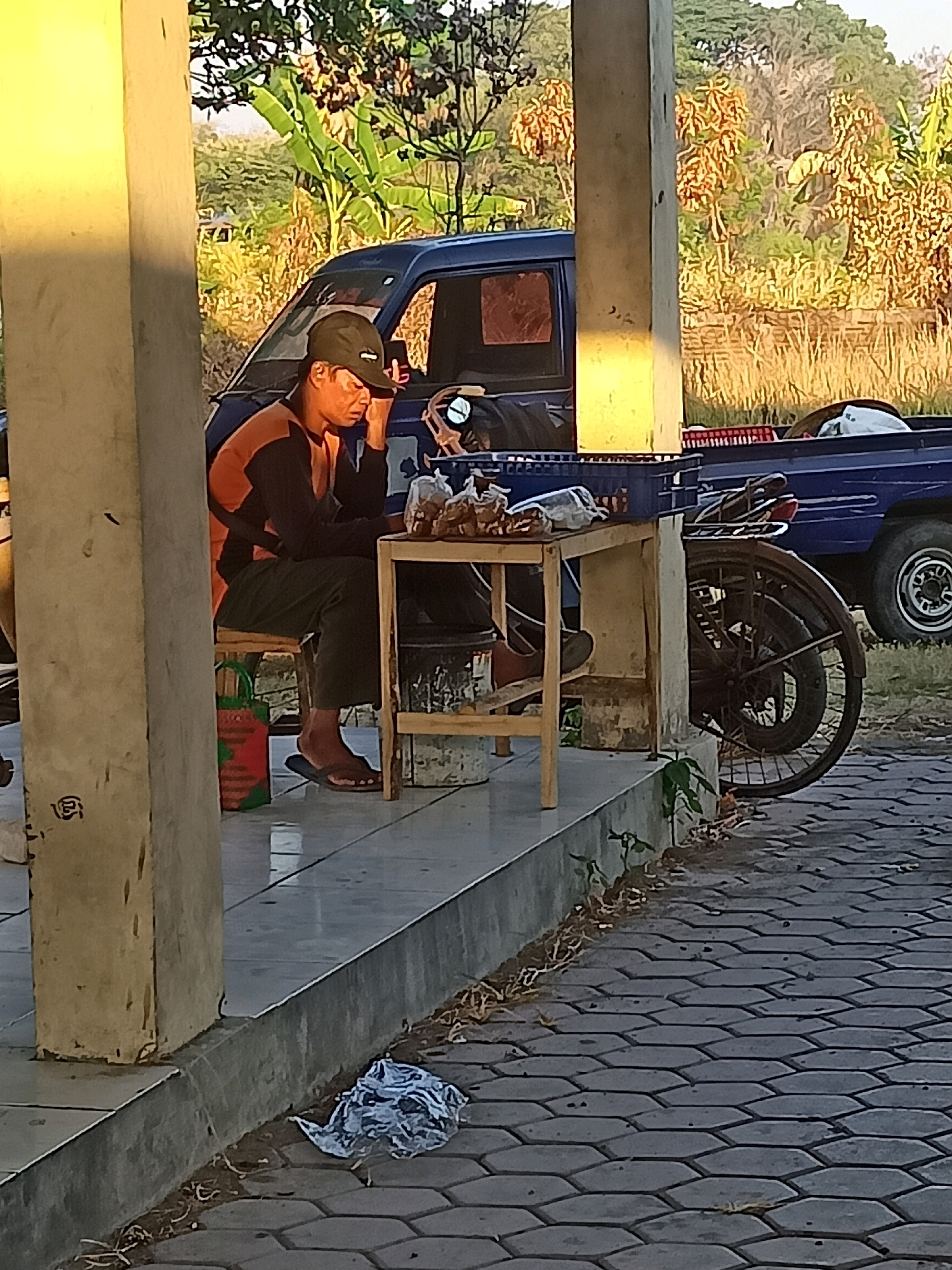 A man in a green cap and an orange and black shirt is sitting on a stool on a concrete porch, leaning on a small wooden table. He is holding a mobile phone to his ear. The table has several bags of food on it. To the right of the table is a bicycle with a large rear wheel and a rack. Behind the man and the table is a blue pickup truck. The porch is next to a paved area with hexagonal stones. In the background, there are trees and foliage. The scene appears to be in a rural or semi-rural area, possibly in Nganti, Indonesia.