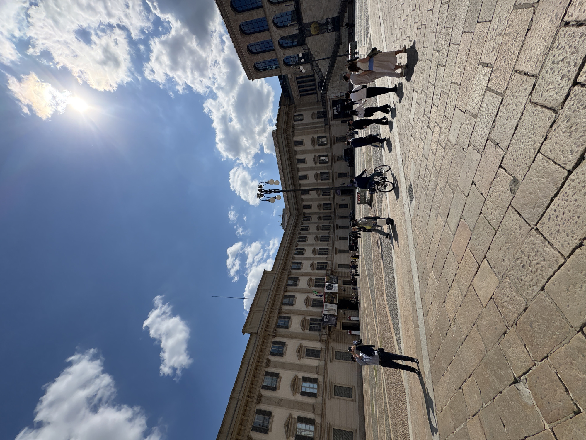 The image shows a street scene in Milan, Italy, with a building facade on the left. A cobblestone square extends to the right. People are present, some walking, and one is on a bicycle. The sky is blue with scattered clouds and the sun is shining. The architecture suggests a historic or government building. Flags are hanging from the facade of the building.