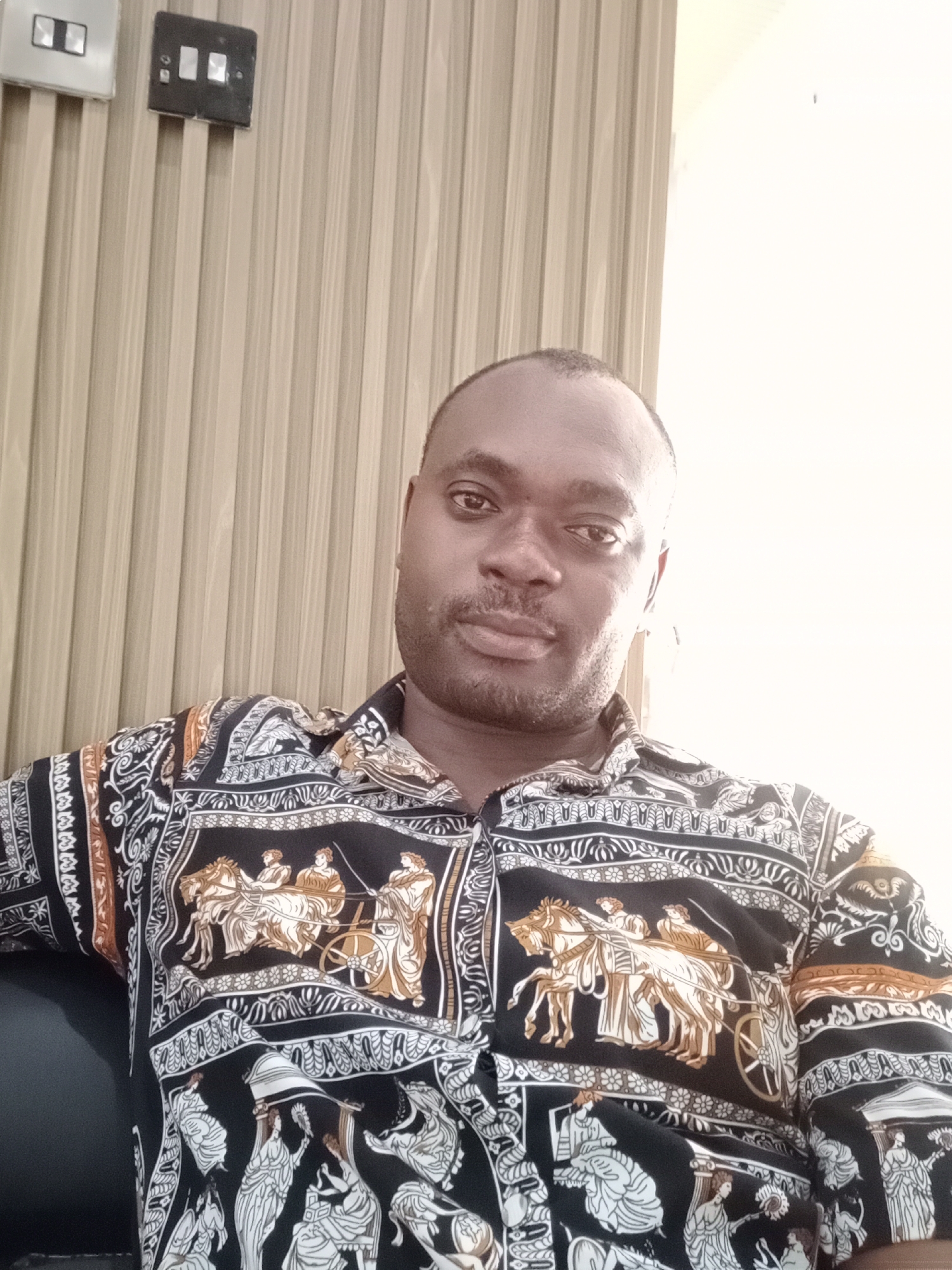 The image depicts a man in an indoor setting. He is sitting, wearing a patterned shirt featuring classical motifs. He appears to be located in Gwarinpa, Nigeria, as indicated by external context. Behind him is a wall with vertical beige panels and a light switch. To the right is a white wall.