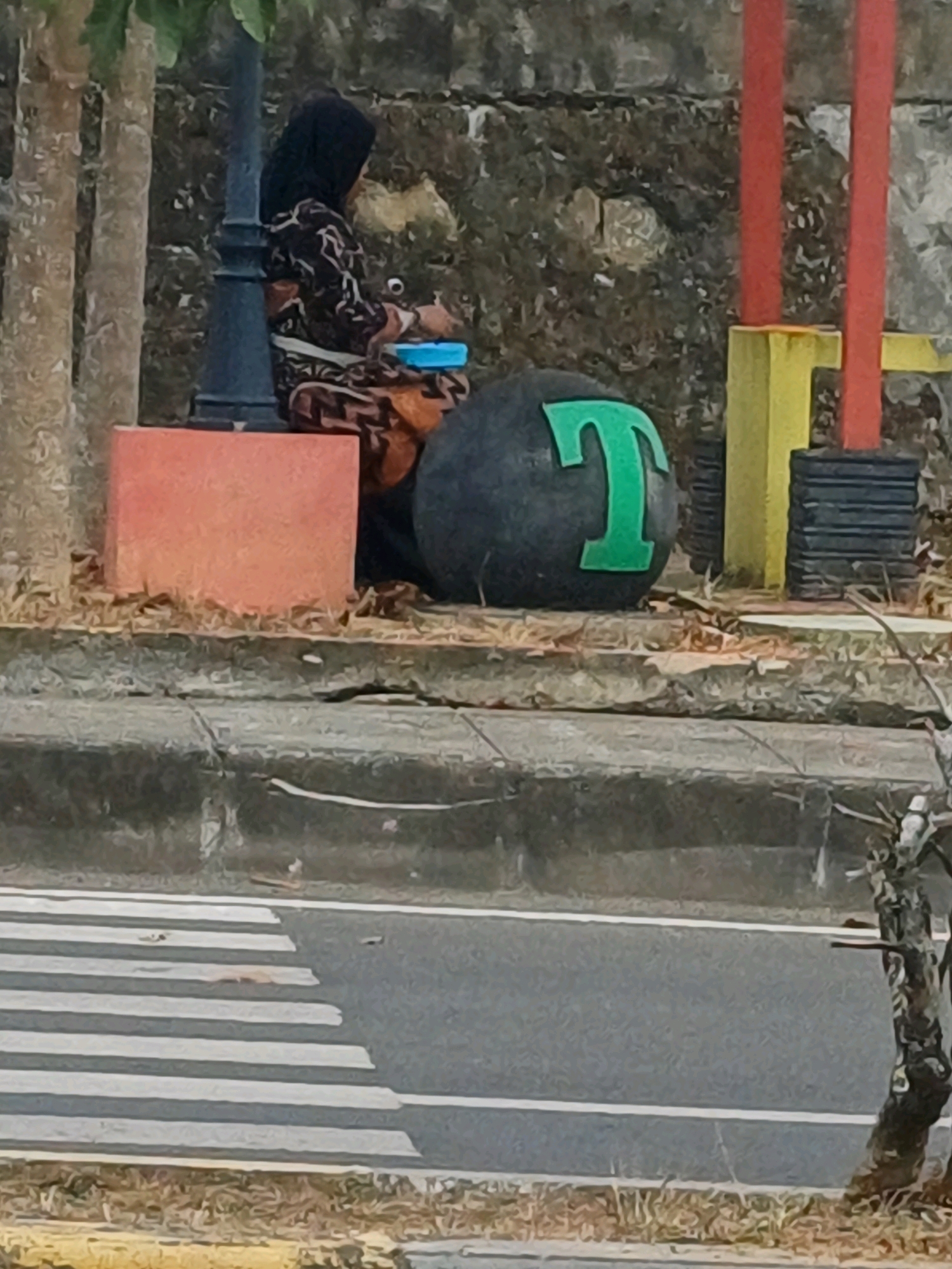 In Pasir Belengkong, Indonesia, a person wearing a headscarf and patterned clothing sits on the ground next to a lamppost. The person is holding a blue object. In front of the person is a large, dark, spherical object with a green "T" symbol on it. Behind them is a textured stone wall. To the right, red posts rise from a yellow base, next to stacked black rectangular objects. A coral-colored block sits to the left of the spherical object. In the foreground, a crosswalk with white stripes is visible on an asphalt road, with dry grass and a tree trunk on the right edge of the frame.