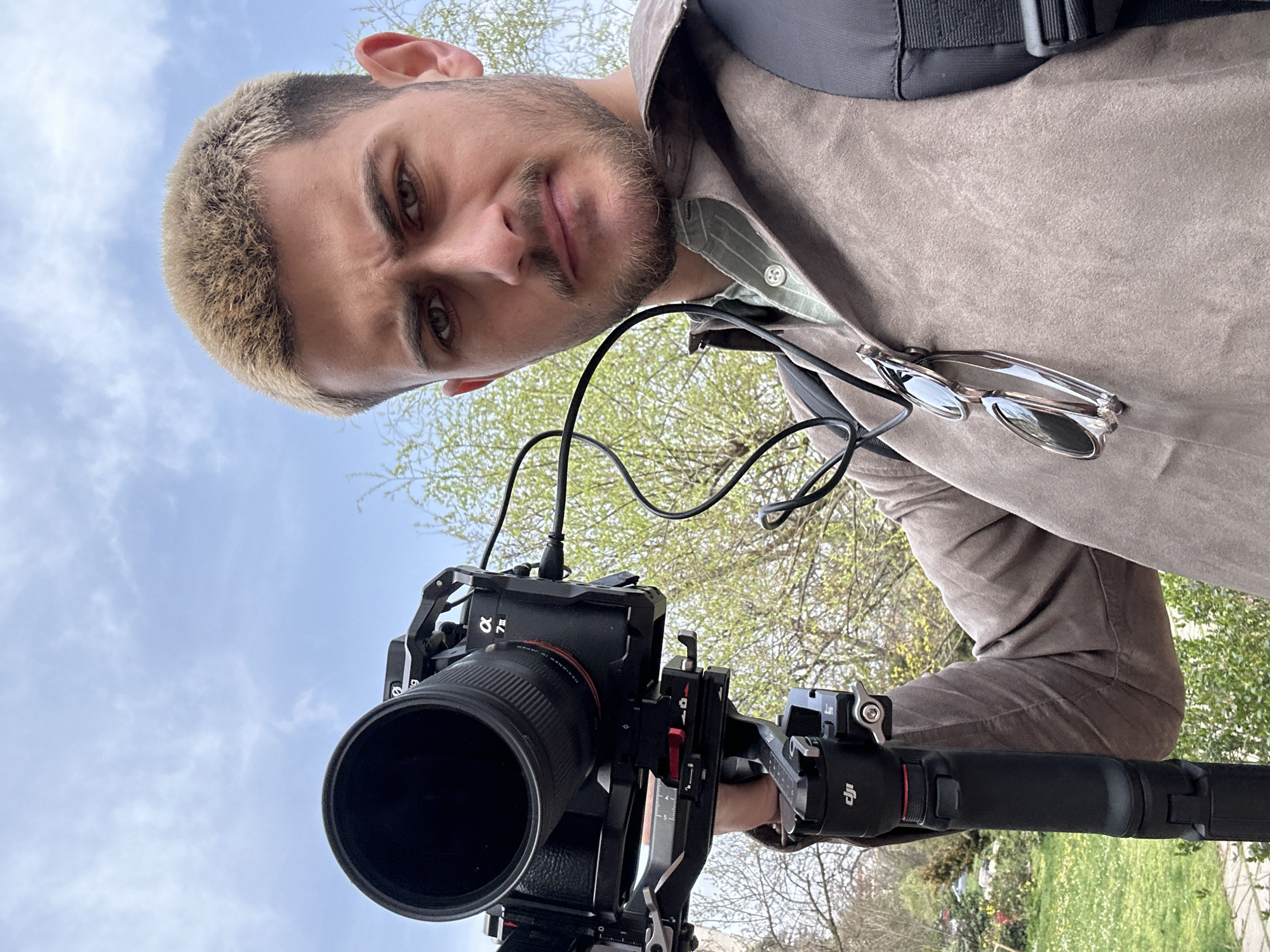 The image is a selfie taken from a low angle, looking up at a man with short, blonde hair and a beard. He is wearing a brown suede jacket over a collared shirt and has sunglasses tucked into his collar. A black camera with a large lens is held in front of him, mounted on a DJI Ronin gimbal. Wires are connected to the camera. In the background, there is a sky with light clouds and trees with green leaves. Given the DJI Ronin gimbal and camera equipment, the man is likely a videographer or photographer.
Without further information, pinpointing the exact location in Sofia, Bulgaria, is not possible. The background suggests a park or area with trees.