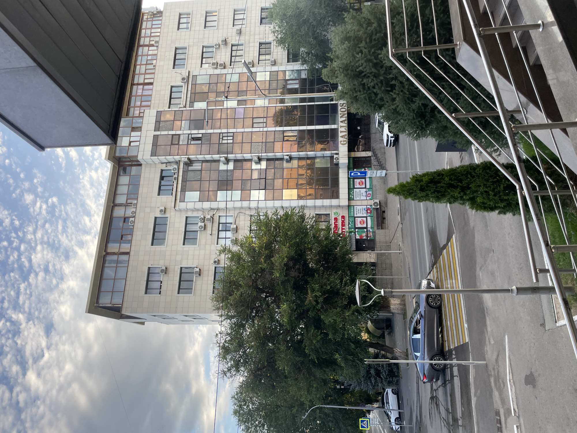 This is a daytime, eye-level shot of a multi-story building with a modern facade. The building has a beige exterior with many windows, some of which are tinted and reflect the sky. The ground floor features storefronts, including a pharmacy with red and green signage. The building is located on a street in Almaty, Kazakhstan, indicated by the Cyrillic text on signs, such as "ГАЛИАНОС" (Galianos) visible on the building's facade and "АПТЕКА" (Apteka) on the pharmacy. A grey sedan is stopped at a pedestrian crossing on the street in front of the building. Several trees, including a large, leafy tree and a tall, slender evergreen, are present. The sky above is partly cloudy. The foreground shows a metal railing and steps leading up to a vantage point from which the photo was taken.