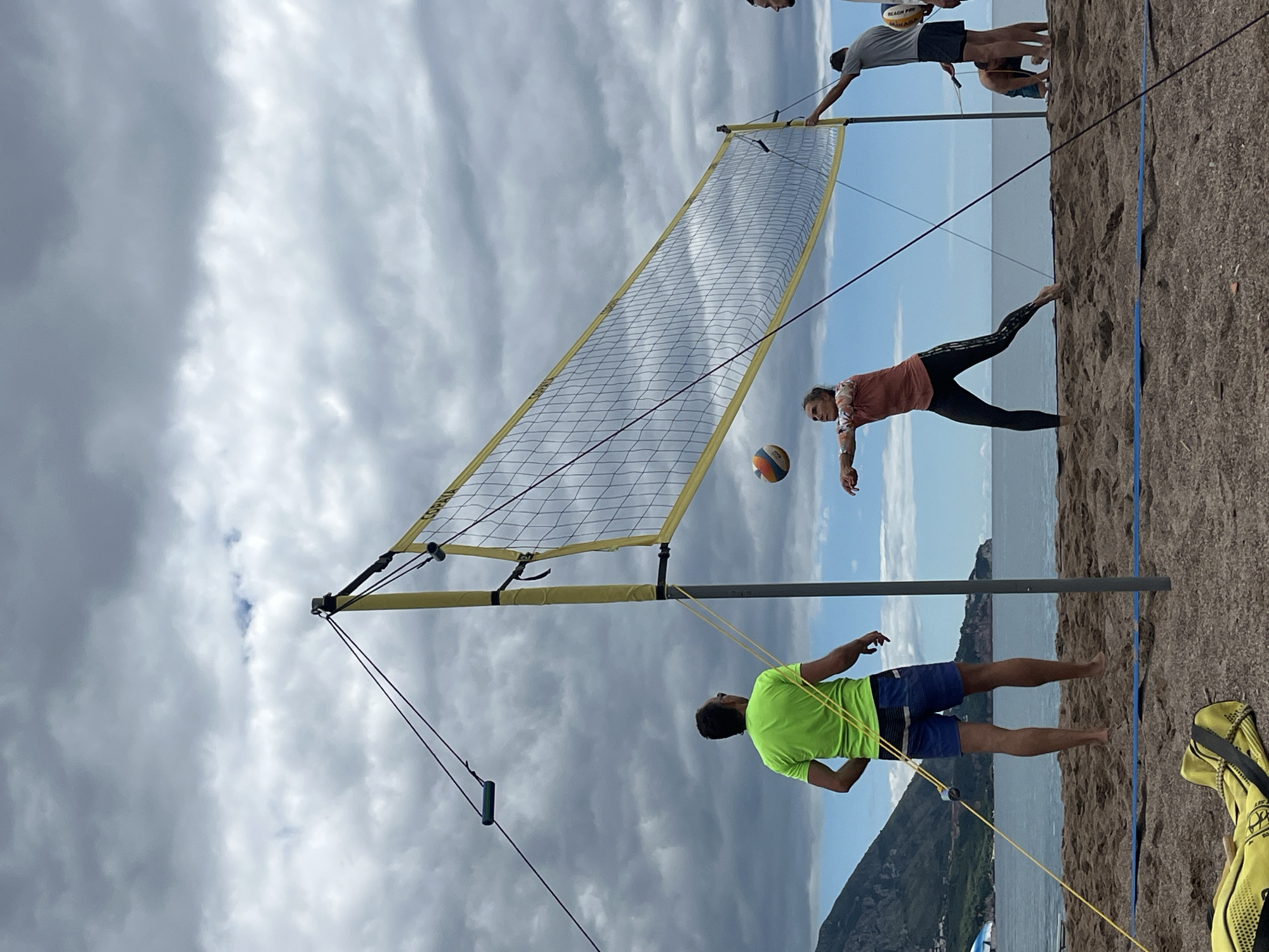 The image depicts a beach volleyball game in progress in Bečići, Montenegro. A beach volleyball net is positioned on a sandy beach with the Adriatic Sea and mountainous terrain in the background. On one side of the net, a woman in a coral-colored top and black leggings is hitting a volleyball. On the opposite side, a man in a bright green shirt and blue shorts is preparing to receive the ball. Other people are visible near the net. The sky is cloudy.