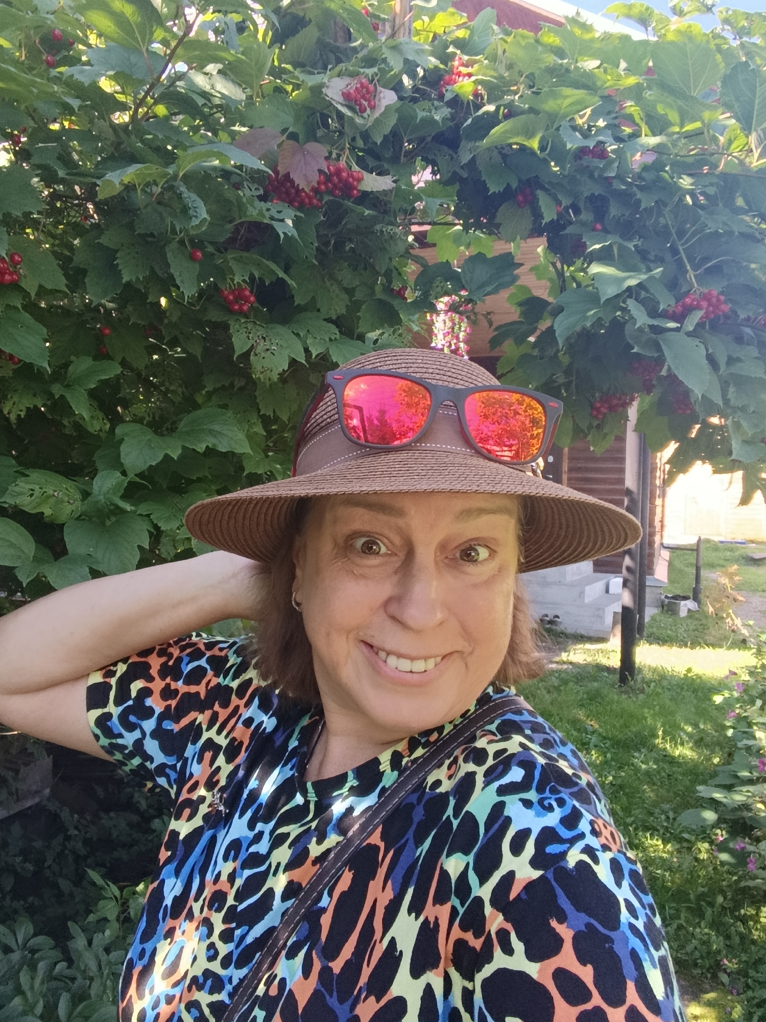 A woman is taking a selfie outdoors in front of a large bush with red berries. She is wearing a straw hat with sunglasses on top and a colorful, leopard-print t-shirt. She has her left arm raised behind her head and is smiling at the camera. The background shows a wooden house and some greenery. The location is likely a residential area in Verea, Russia.
