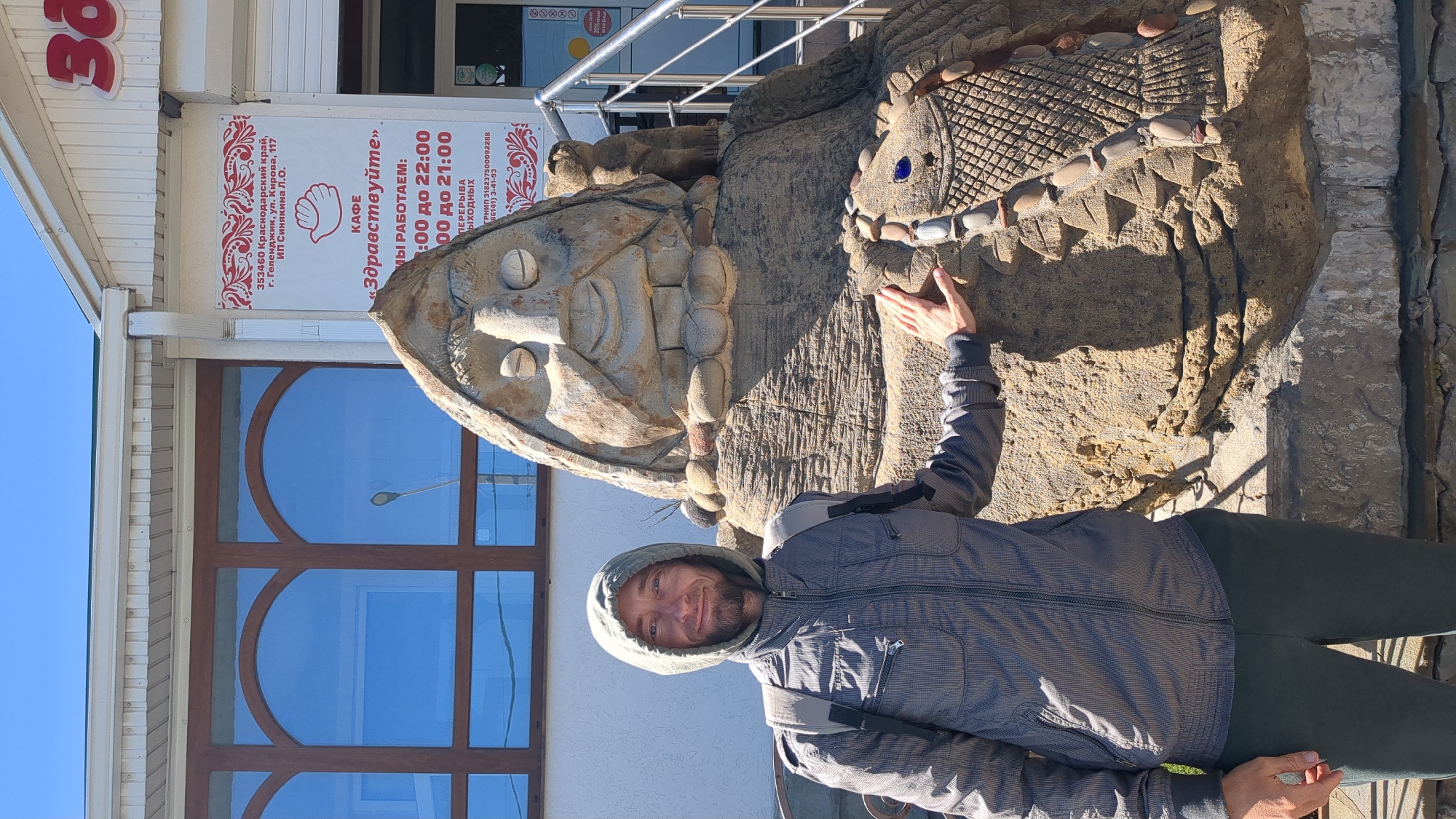 A man is depicted standing next to a large, ornate stone sculpture under a clear blue sky. He wears a gray hooded jacket and dark trousers, looking at the camera with a friendly smile, and has his right hand resting on the sculpture.
The sculpture is a prominent feature, carved from stone or concrete, resembling an ancient or tribal-style anthropomorphic figure. It features a detailed, smiling face with large eyes and a textured headdress. The body of the sculpture is heavily textured, adorned with embedded stones or shell-like elements, and blue glass beads are visible on its side.
In the background, a white building with distinctive arched windows is visible. The bright sunlight indicates it is daytime, casting clear shadows.
A sign on the building, written in Russian, provides details about the location and business. It states the address as "353460 Краснодарский край, г. Геленджик, ул. Кирова, 117 ИП Синякина Л.О." (353460 Krasnodar Krai, Gelendzhik, Kirova St., 117 IP Sinyakina L.O.). The establishment is identified as "КАФЕ «Здравствуйте»" (CAFE "Zdravstvuyte" or "Hello"). Partial working hours are displayed, indicating operation until 22:00 and 21:00, with an incomplete mention of "ПЕРЕРЫВА ВЫХОДНЫХ" (Break, Weekends). A registration number (ГРНИП 318237500092288) and a phone number (86141) 3-41-93) are also visible. The image captures a cheerful moment in Gelendzhik, Russia.