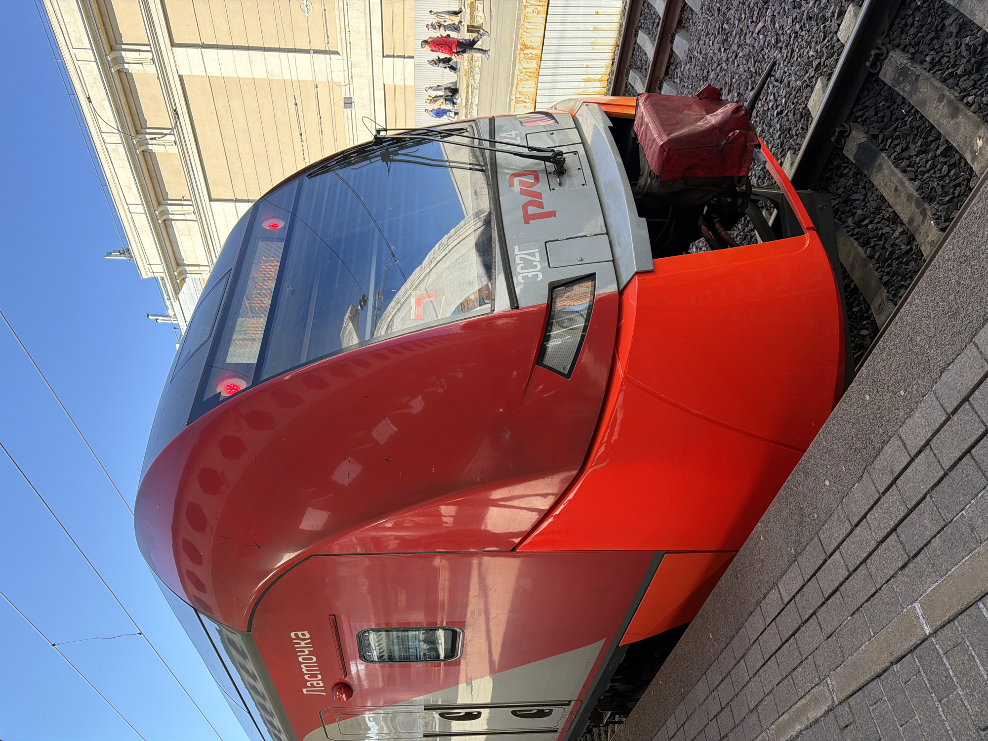 A red train sits at a station platform, facing the right side of the frame. The train car is marked with Cyrillic text. Above the train is a concrete building with a blue sky in the upper-left corner of the frame. People stand in the background on the platform. To the right of the train are railroad tracks. The station is most likely located in the city of Saint Petersburg, Russia.