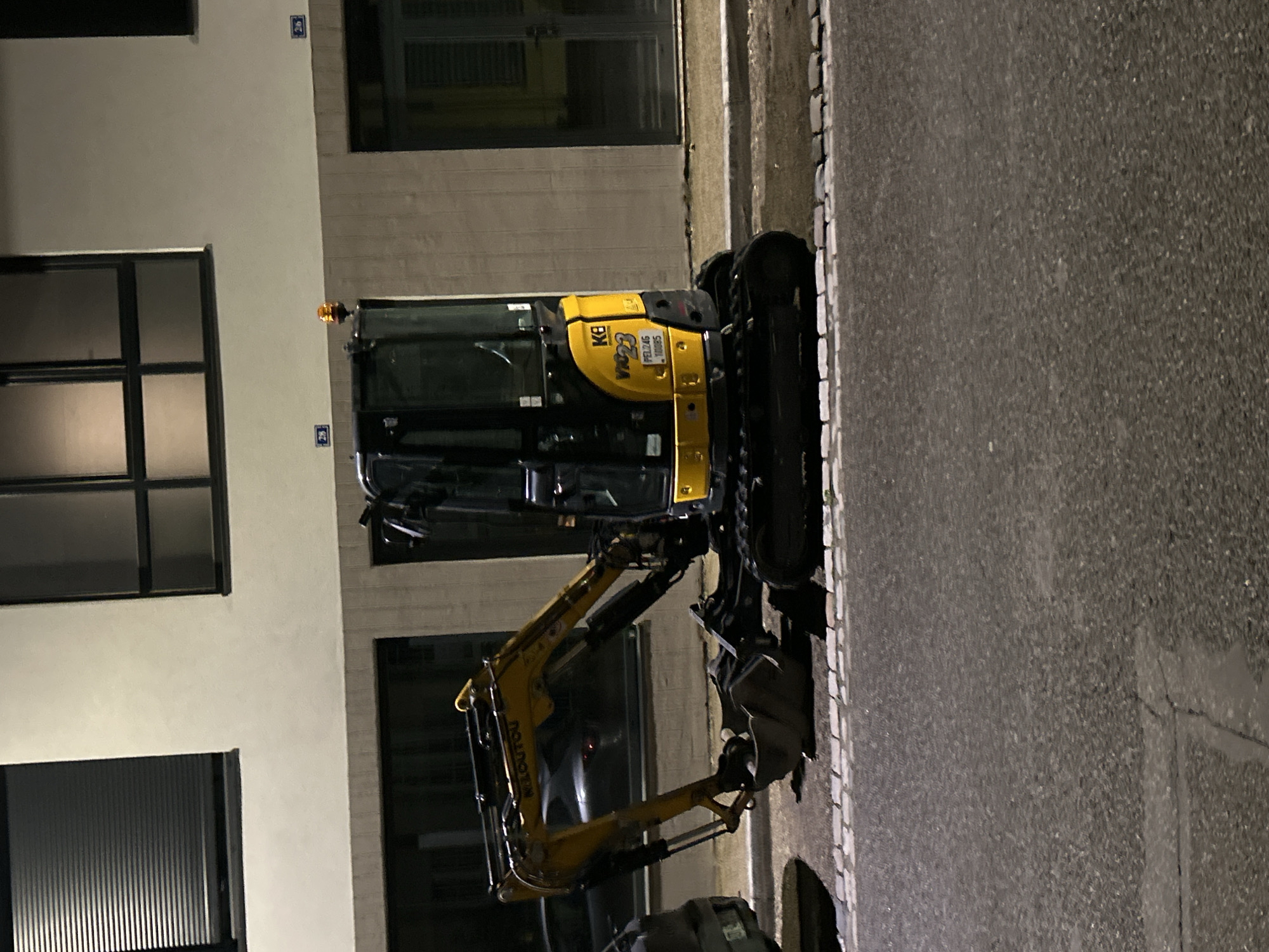 The image depicts a Komatsu PC26MR-3 excavator parked on the side of a street. The excavator is yellow and black, with a bucket attachment. The background includes a building with several windows. Based on the street number visible on the building facade, the location is Strasbourg, France. The scene appears to be at night due to the darkness and artificial lighting. The street is paved. There are no people visible in the image.