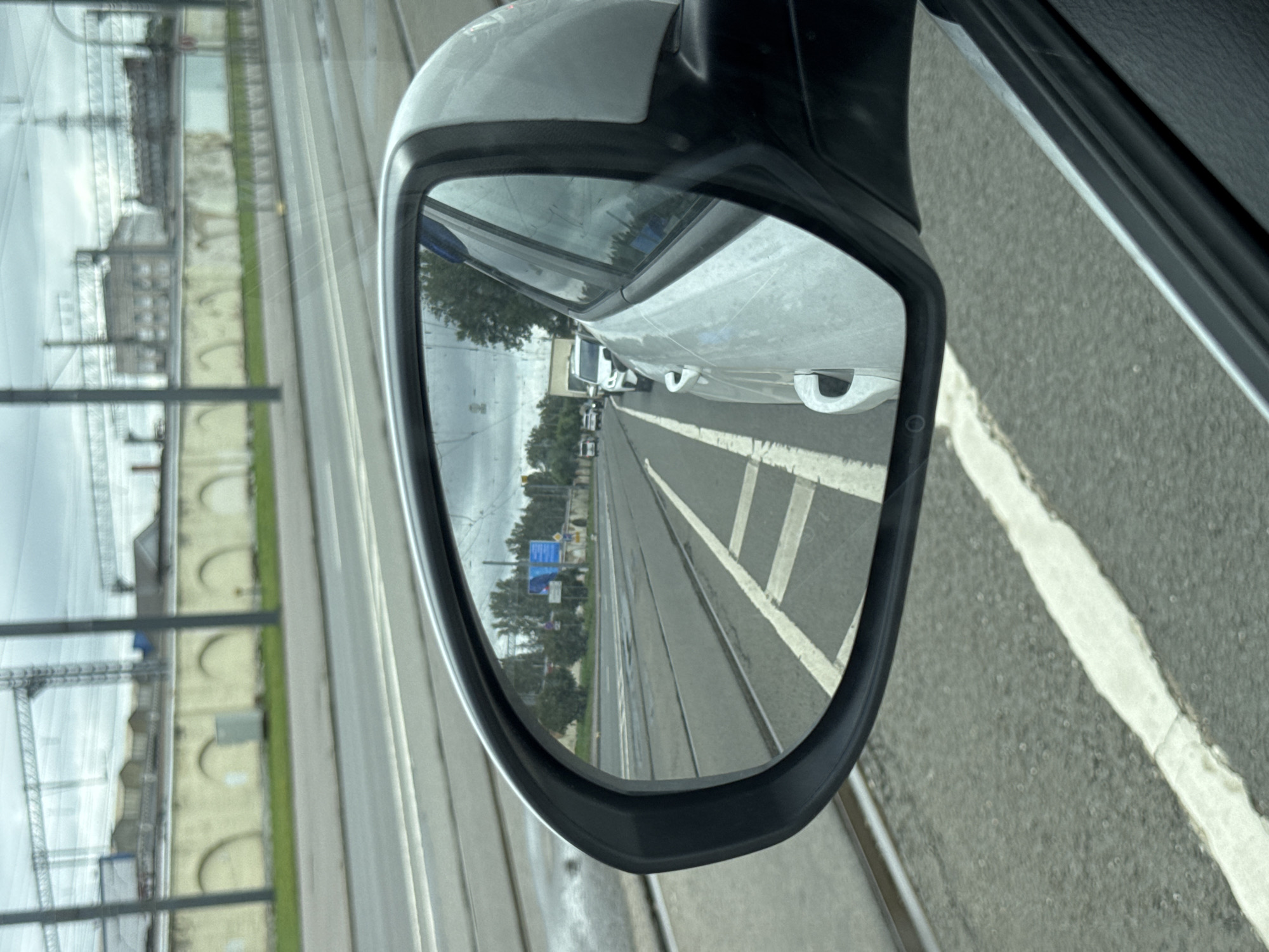 The image shows a view through a car's side mirror, reflecting a street scene in Saint Petersburg, Russia. The street appears to have tram tracks running along it, with cars stopped in traffic behind. In the distance, there is a long, low building with arched openings, possibly a historical structure or industrial facility, and overhead electrical lines for trains or trams are visible. The sky is overcast. The car itself is white and parked next to the tram tracks.