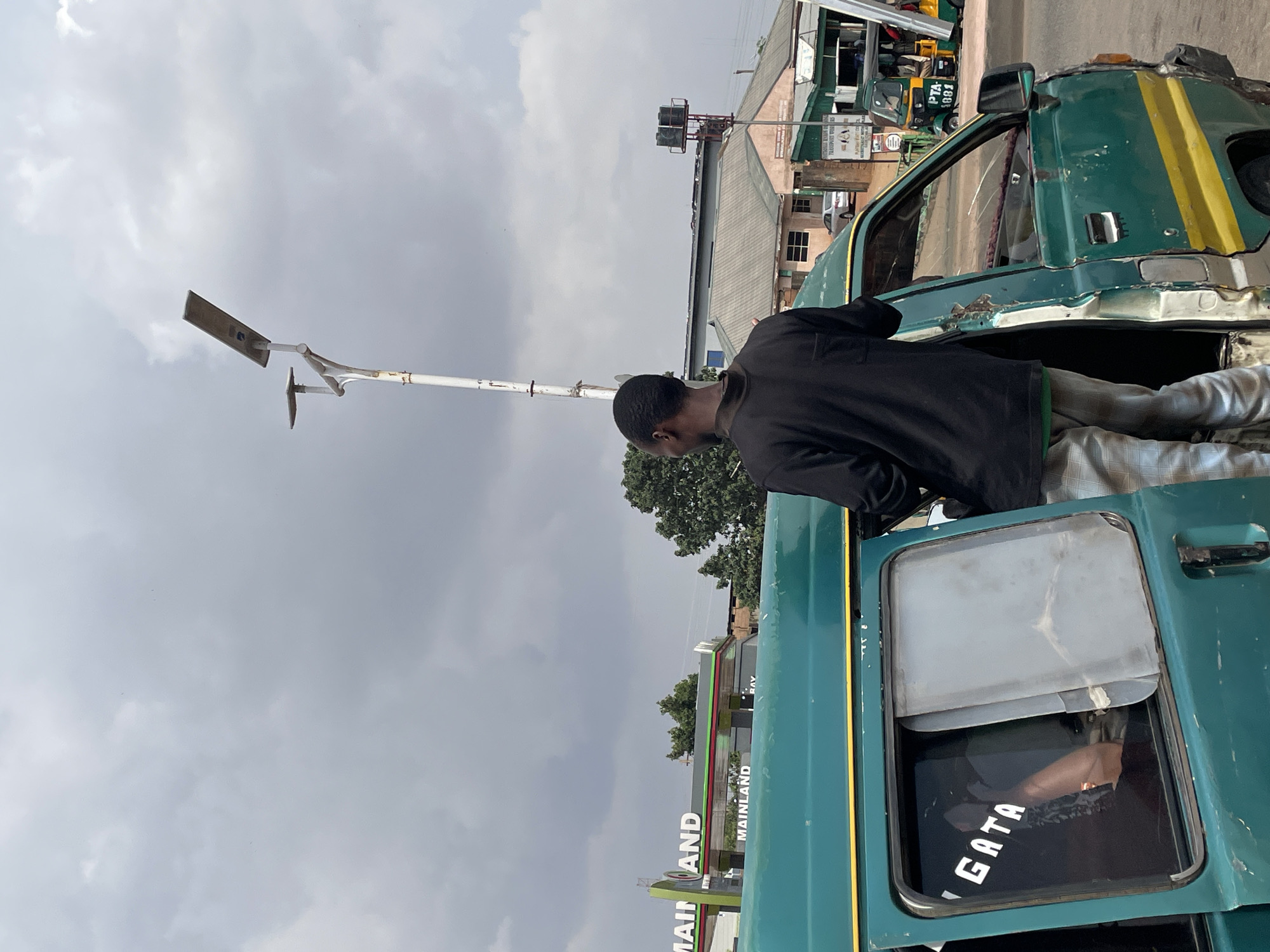 A medium-level, outdoor shot depicts a man standing in the open doorway of a green vehicle. He is wearing a black long-sleeved shirt and light-colored pants. The vehicle appears to be a commercial transport vehicle, with writing visible on the back door. A lamppost is visible nearby. The scene is set in Jos, Nigeria, as indicated by a sign in the background. A commercial transport vehicle and building can be seen in the background. The sky is overcast.
