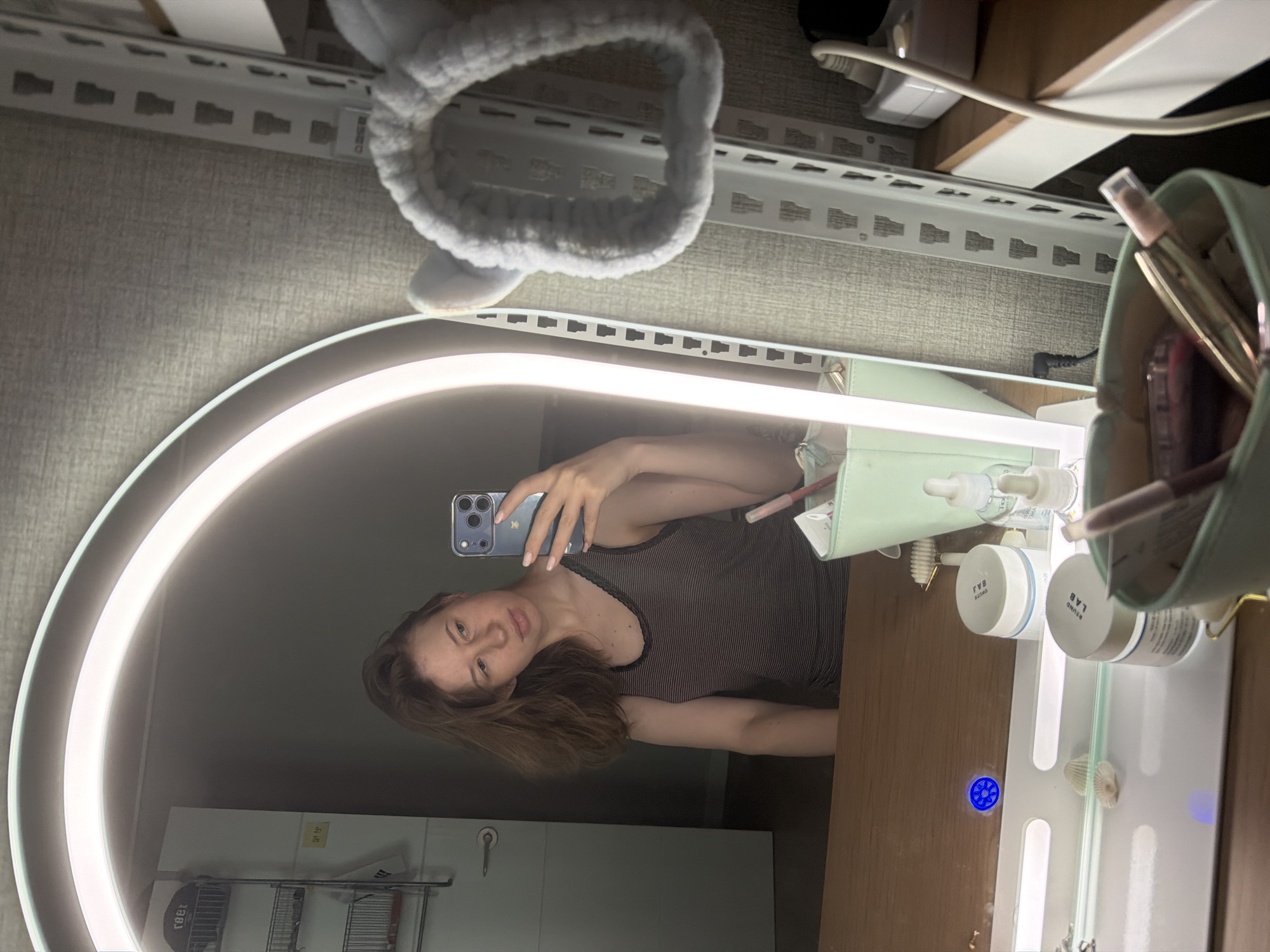 A young woman with shoulder-length brown hair is taking a selfie in a mirror. She is wearing a dark striped tank top and has light-colored nail polish on her fingernails. She is holding a smartphone in her right hand, which is positioned to capture her reflection. The mirror is illuminated by a bright ring light around its edge.
In the reflection, the woman is seated at a wooden vanity. On the vanity are several cosmetic products, including jars and bottles, along with some seashells. A light green cosmetic organizer holding tubes and bottles is visible in the foreground of the vanity. Behind the vanity, a white metal shelf unit is present, and a light gray, fuzzy headband with ear-like accents rests on it. The background on the right side of the mirror shows a textured wall and a white power outlet with a black cable plugged in. The background on the left side of the mirror shows a white door with a silver handle and a shelf with items on it.
The scene appears to be a personal vanity area, likely in a home, possibly in Dongducheon-si, South Korea, given the information provided.
