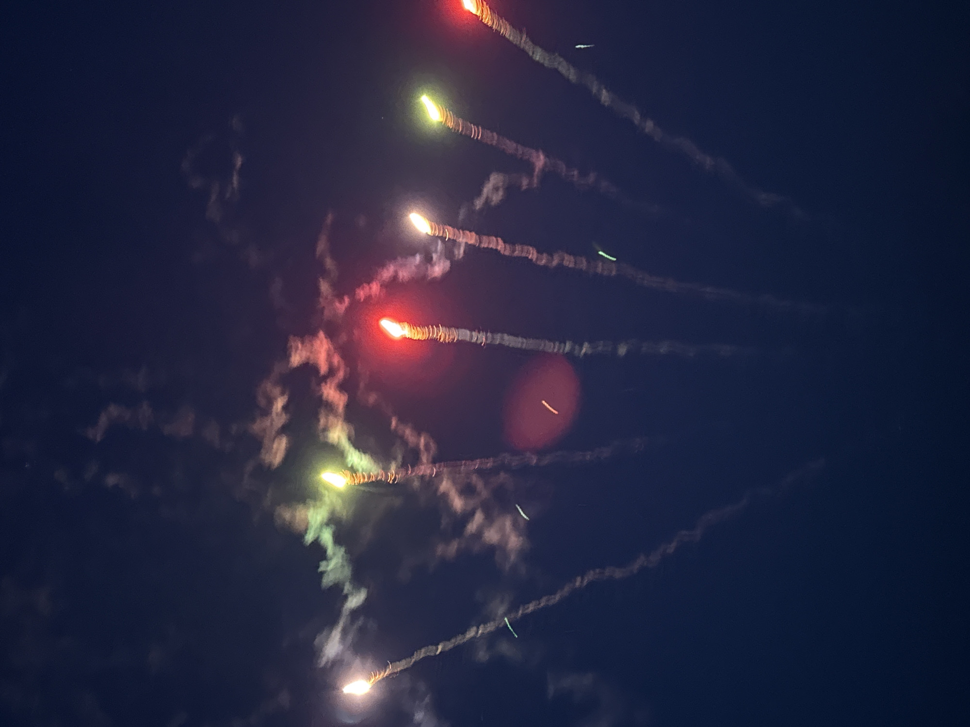 The image depicts multiple fireworks streaking across a dark night sky. The fireworks exhibit varying colors including red, orange, yellow, and green, leaving trails of smoke and light in their wake. Some smoke plumes spread and diffuse into cloud-like formations. The location of the image is not identifiable from the image itself.