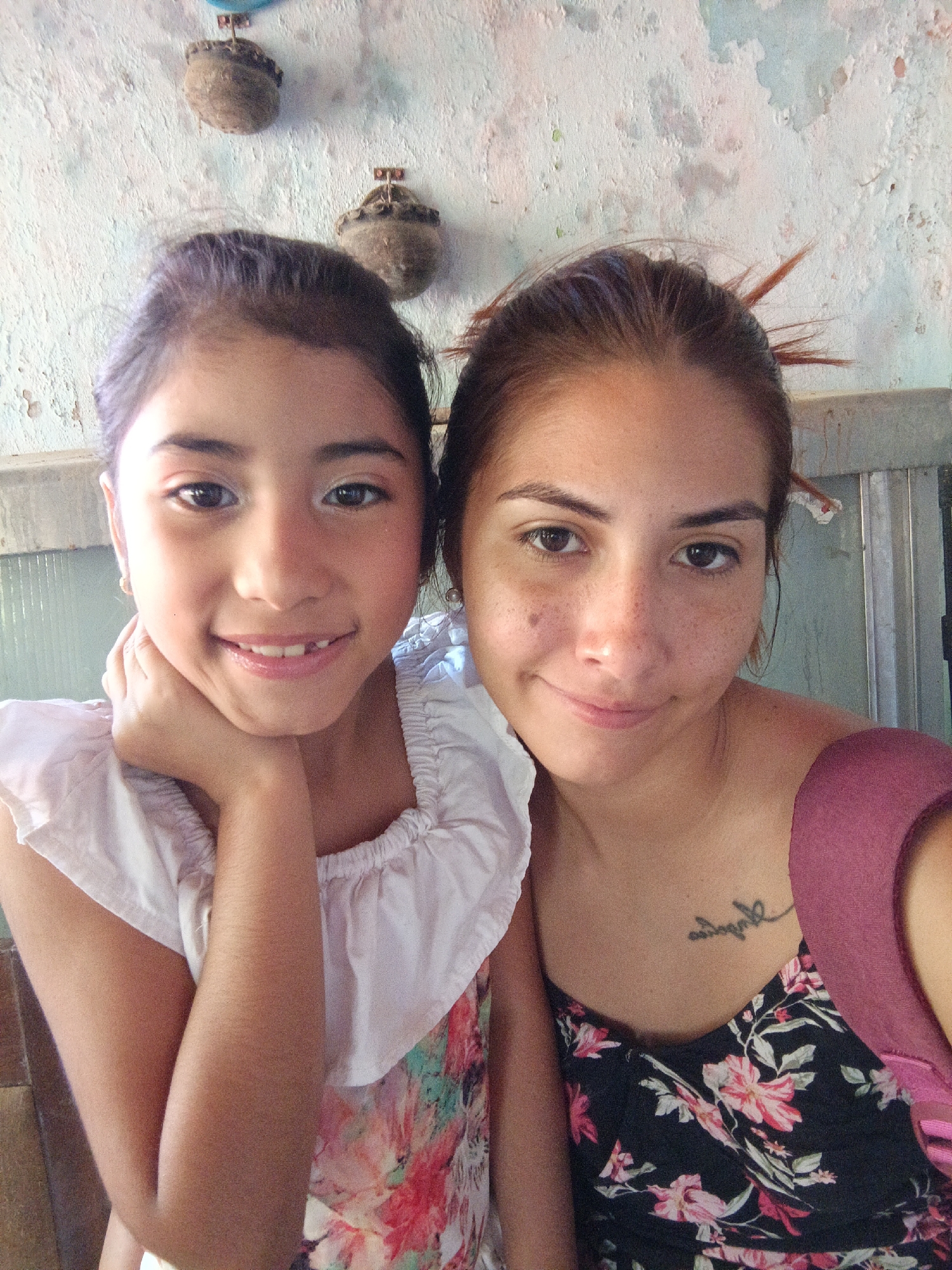 Two females are posing for a selfie. The female on the left is younger, approximately 10 years old, with brown hair and dark eyes. She has a light complexion and is wearing a white and floral print top. Her head is tilted to the left, and her left hand is supporting her chin. The female on the right appears to be an adult, likely in her 20s. She has brown hair pulled back, brown eyes, and freckles. She is wearing a floral print top with a purple strap. She has a small tattoo on her upper chest, just above the neckline of her dress. The background is a textured wall with two small, possibly decorative, items hanging on it. There is a metal grate or door visible in the upper right corner of the frame. The image appears to be taken in Juan Griego, Venezuela.