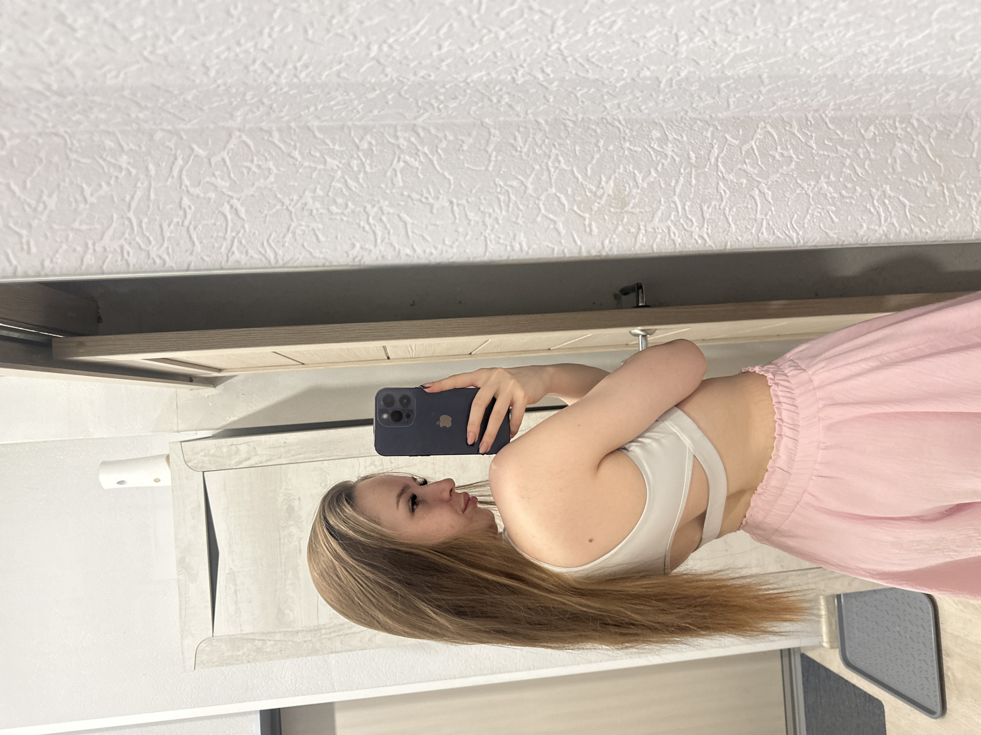 A young woman with long blonde hair is holding a dark blue smartphone in her right hand, taking a selfie in an indoor setting. She is wearing a white sports bra and pink pants. The background includes a textured white ceiling, a light wood-paneled section, and a white cabinet with a light gray countertop area. The location is Unknown, Unknown.