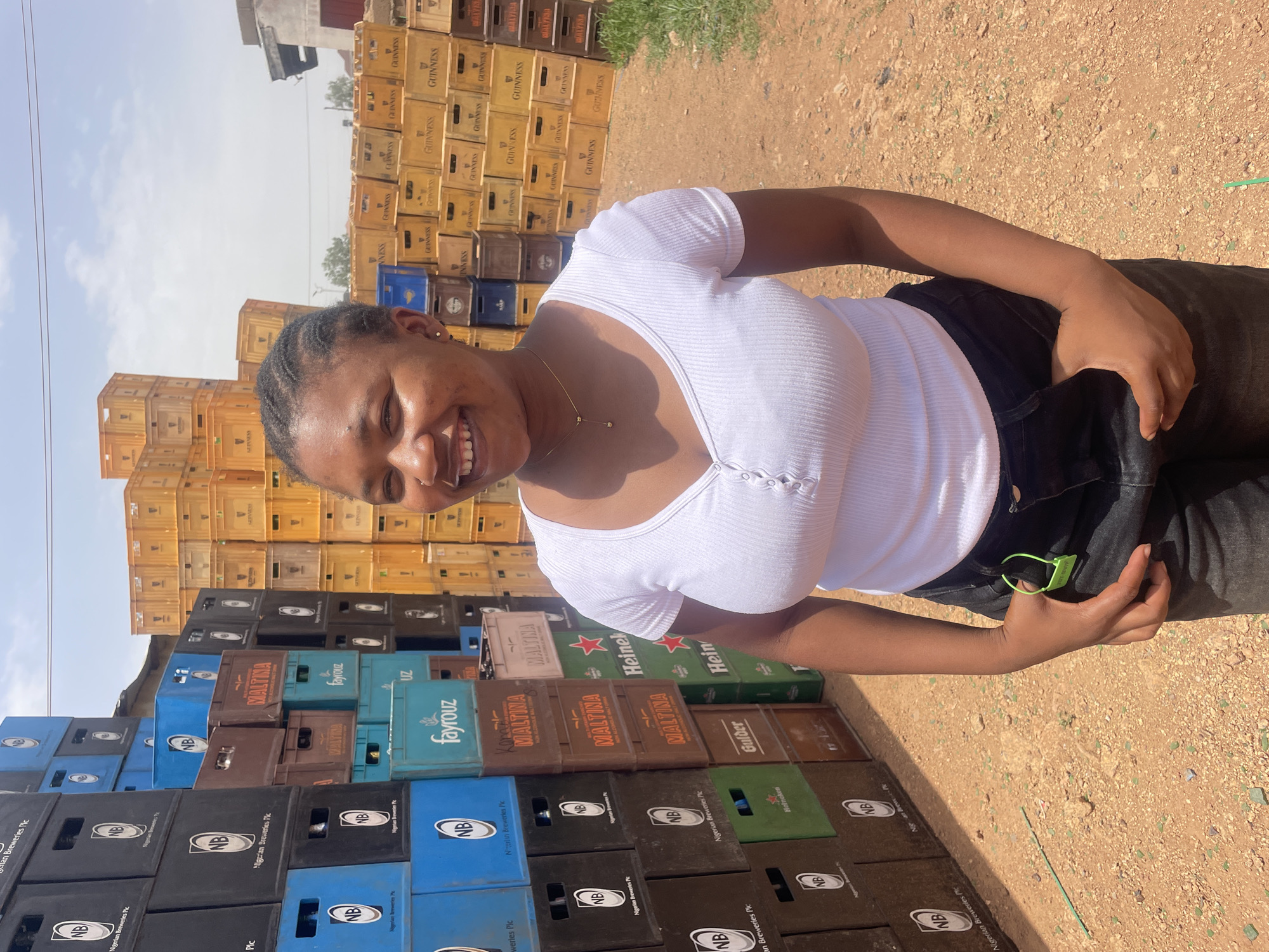A woman stands smiling in front of stacks of beverage crates. The crates are in varying colors: blue, green, brown, yellow, and black, and feature brands such as Guinness, Maltina, Heineken, and Fayrouz. The ground is a dusty brown. A tag is visible on the woman's pants. The woman is wearing a white shirt and black pants. The location is Gura Top, Nigeria.