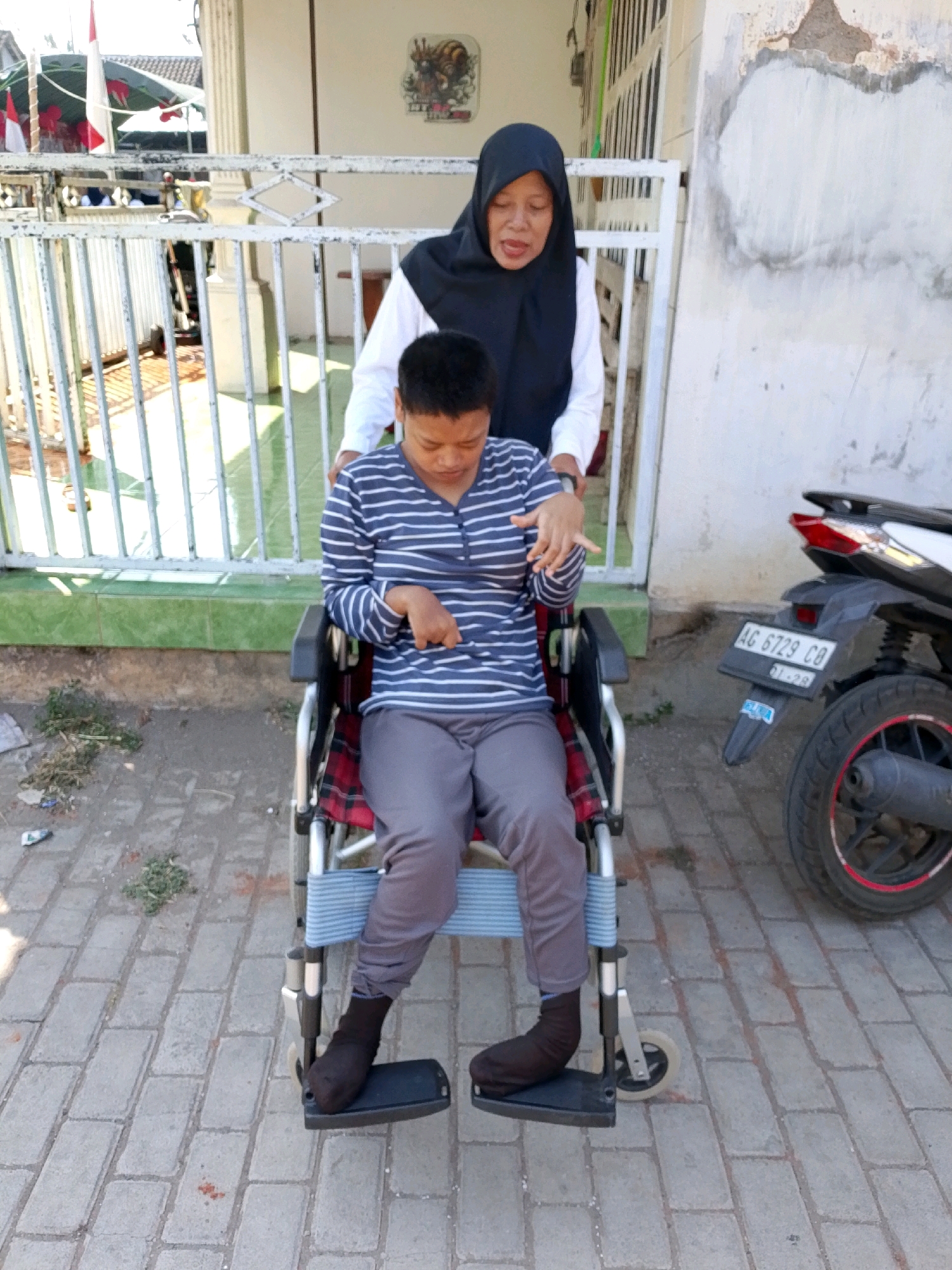 A woman wearing a black hijab and a white shirt is pushing a boy in a wheelchair. The boy is wearing a blue and white striped shirt and grey pants. They are on a paved pathway in front of a building. A motorcycle is parked to the right of the frame. The location is identified as Banyakan, Indonesia.
