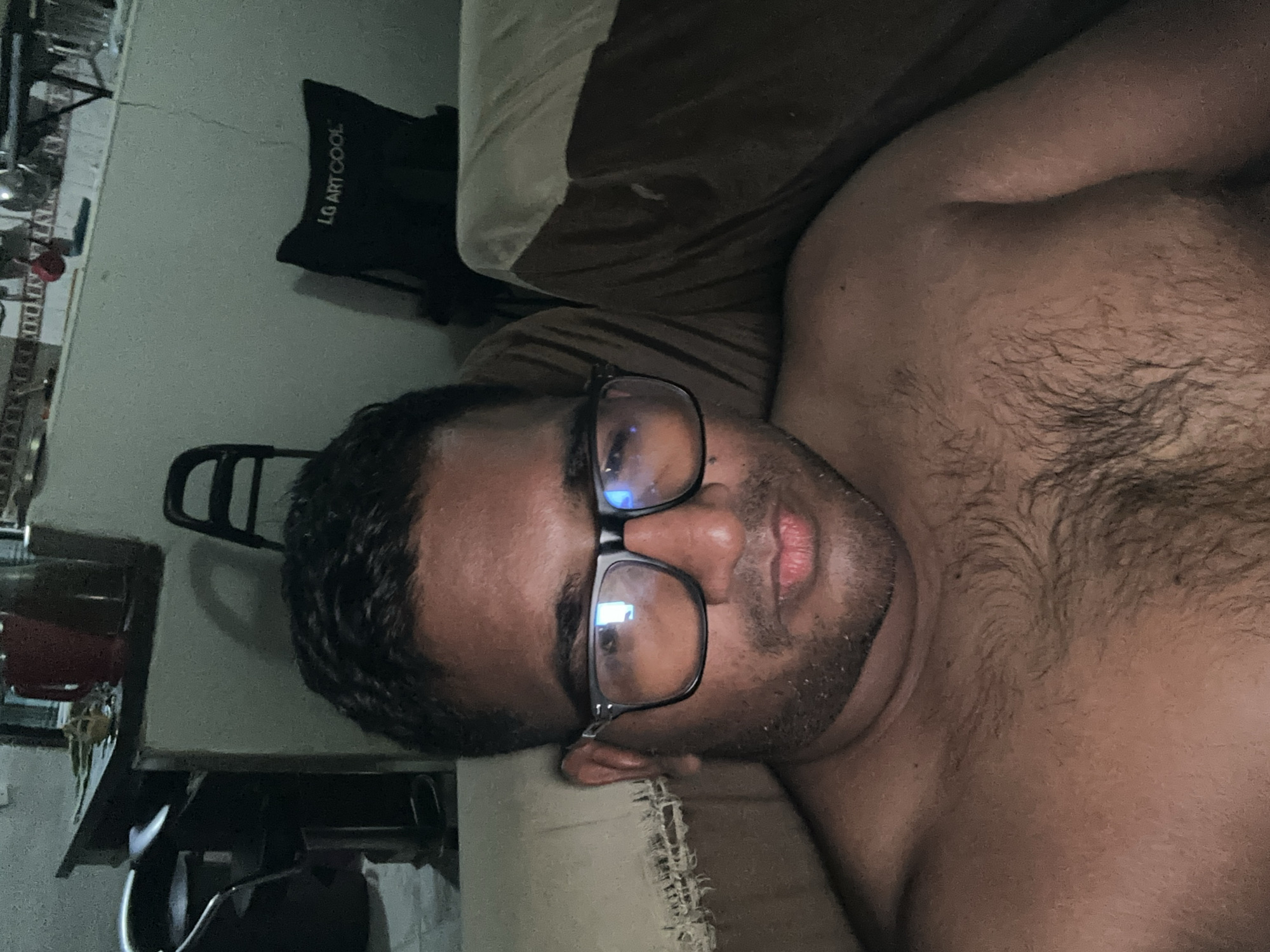 A man with dark hair and glasses lies on a light-colored cushion. His upper torso is exposed. The location is Parroquia Ocumare, Venezuela, though specific landmarks or features confirming this are absent in the image. An "LG ART COOL" branded object is visible in the background. A shelf and various objects are also visible in the periphery.