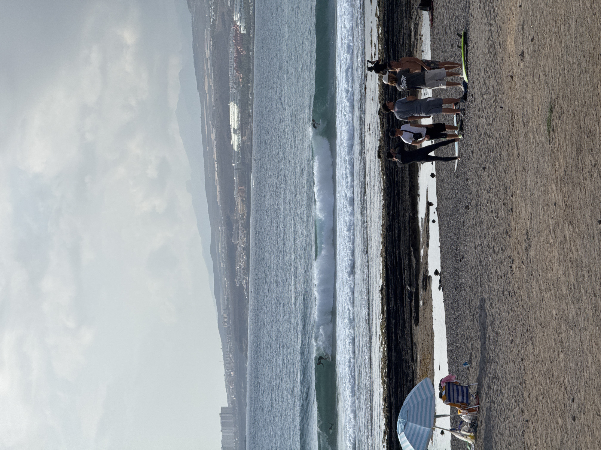 In Arona, Spain, a group of surfers stands on a rocky shore next to a sandy beach, with the ocean in the background. The sky is overcast, with clouds overhead and a hazy view of distant mountains and buildings on the horizon. The water is choppy, with a large wave breaking, and two surfers are visible riding it. On the beach, a blue and white striped umbrella provides shade over a beach chair and some belongings. The group of surfers on the shore includes at least five individuals, some wearing wetsuits, and several surfboards are placed on the ground.