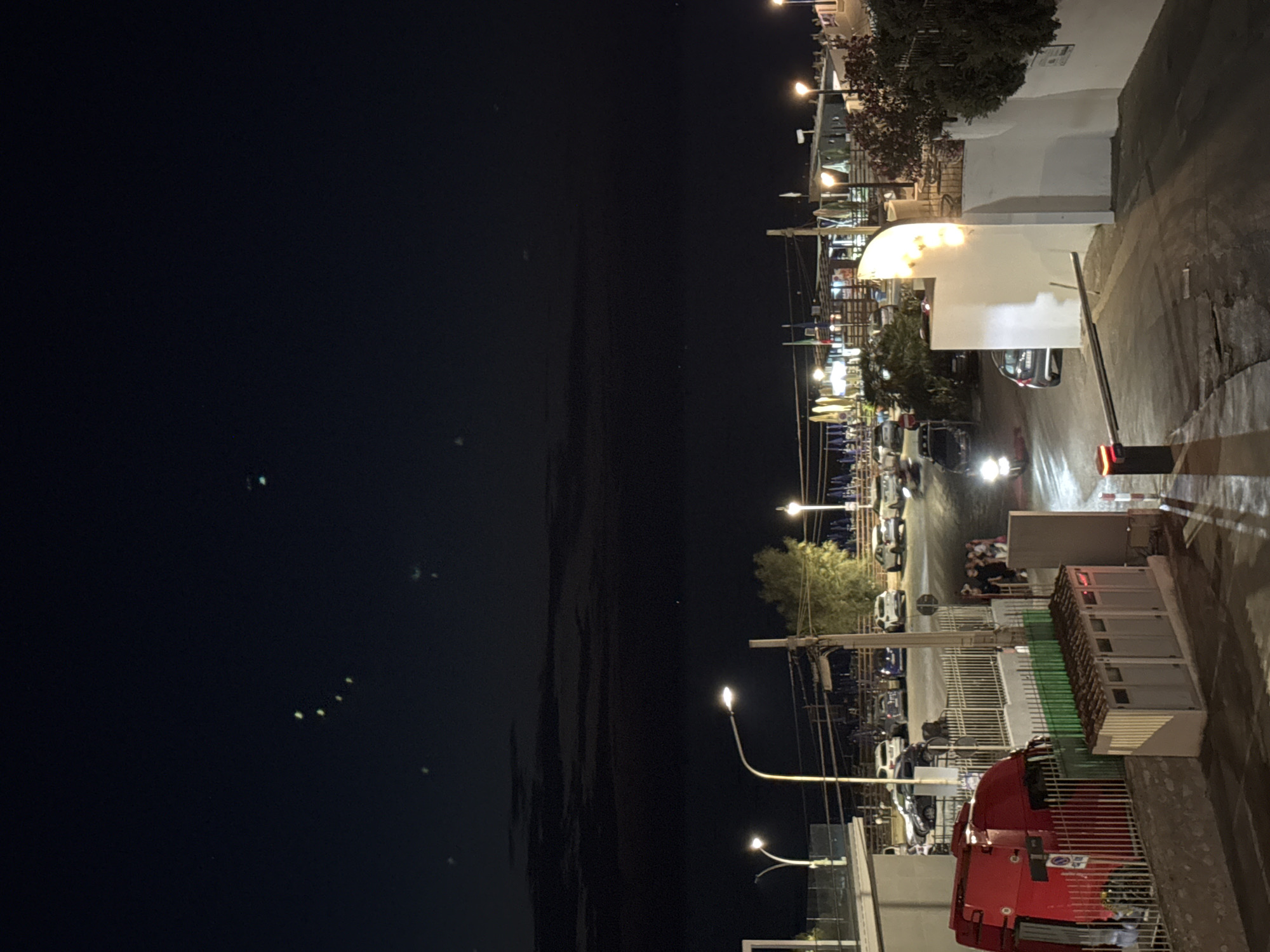 The image shows a street scene at night in Capaci, Italy. A red truck is parked on the left side of the frame. Across the street, there is a parking lot with several cars, and a building with lights and umbrellas in front of it. People are visible walking near the building. There are streetlights illuminating the area, and the sky is dark with some faint lights visible. The environment appears to be a coastal town, with beach umbrellas suggesting proximity to the sea. A barrier gate is visible on the right side of the road.