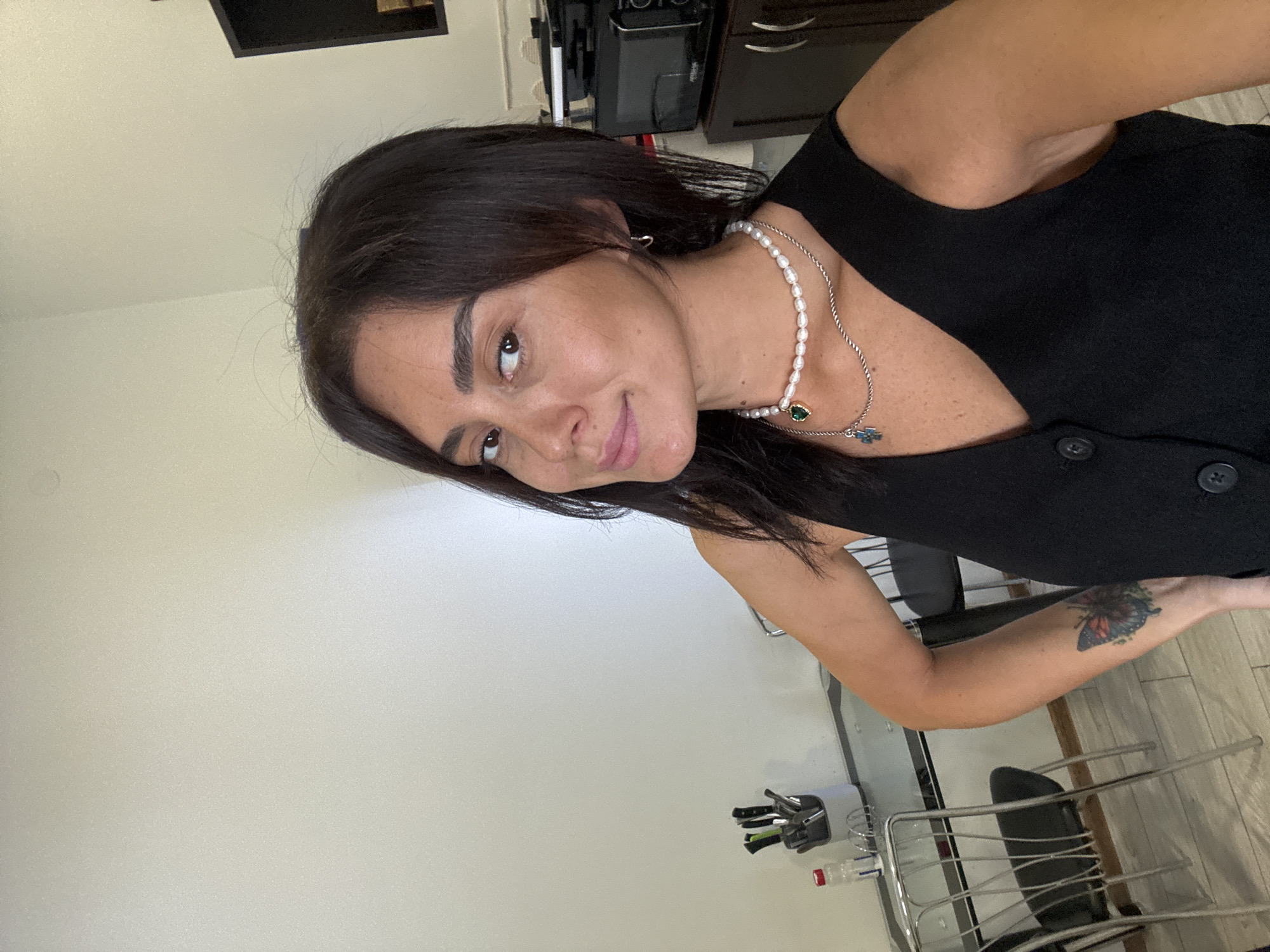 A woman with dark hair and brown eyes is taking a selfie in what appears to be a domestic setting, possibly an apartment kitchen in Tbilisi, Georgia. She is wearing a black sleeveless vest and two necklaces, one with pearls and a green pendant, and another with a silver chain and a blue pendant. On her left forearm, there is a tattoo of a colorful butterfly. In the background, a glass table with chairs, a knife block with utensils, and a water bottle are visible. Further back, a dark wooden cabinet with a black appliance on top is present. The walls are painted a light neutral color.