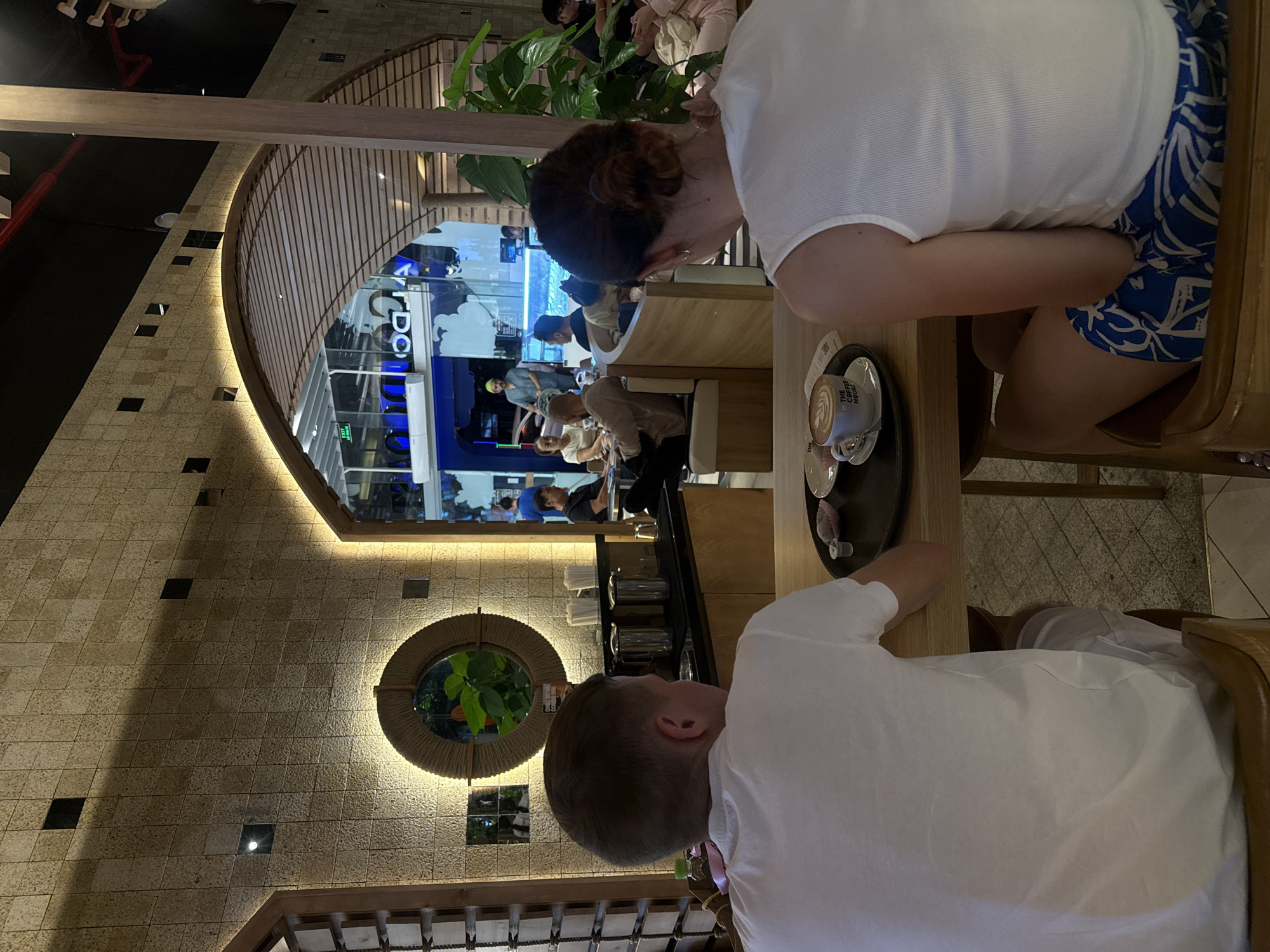The image shows a cafe interior in Nha Trang Ward, Vietnam. Two individuals, a man and a woman, are seated at a table in the foreground, facing away from the camera. The man is wearing a white t-shirt, and the woman is wearing a white tank top and patterned shorts. On the table in front of them is a coffee cup with latte art, a small dish with a dessert, and a coaster.
In the background, other patrons are visible seated at tables and on benches. The cafe features a textured stone wall, a large arched opening with a wooden slatted ceiling, and ambient lighting. Beyond the arched opening, a brightly lit space with blue accents and what appears to be an aquarium or water feature is visible. Green plants are also present in the environment. The overall atmosphere appears to be casual and relaxed.
