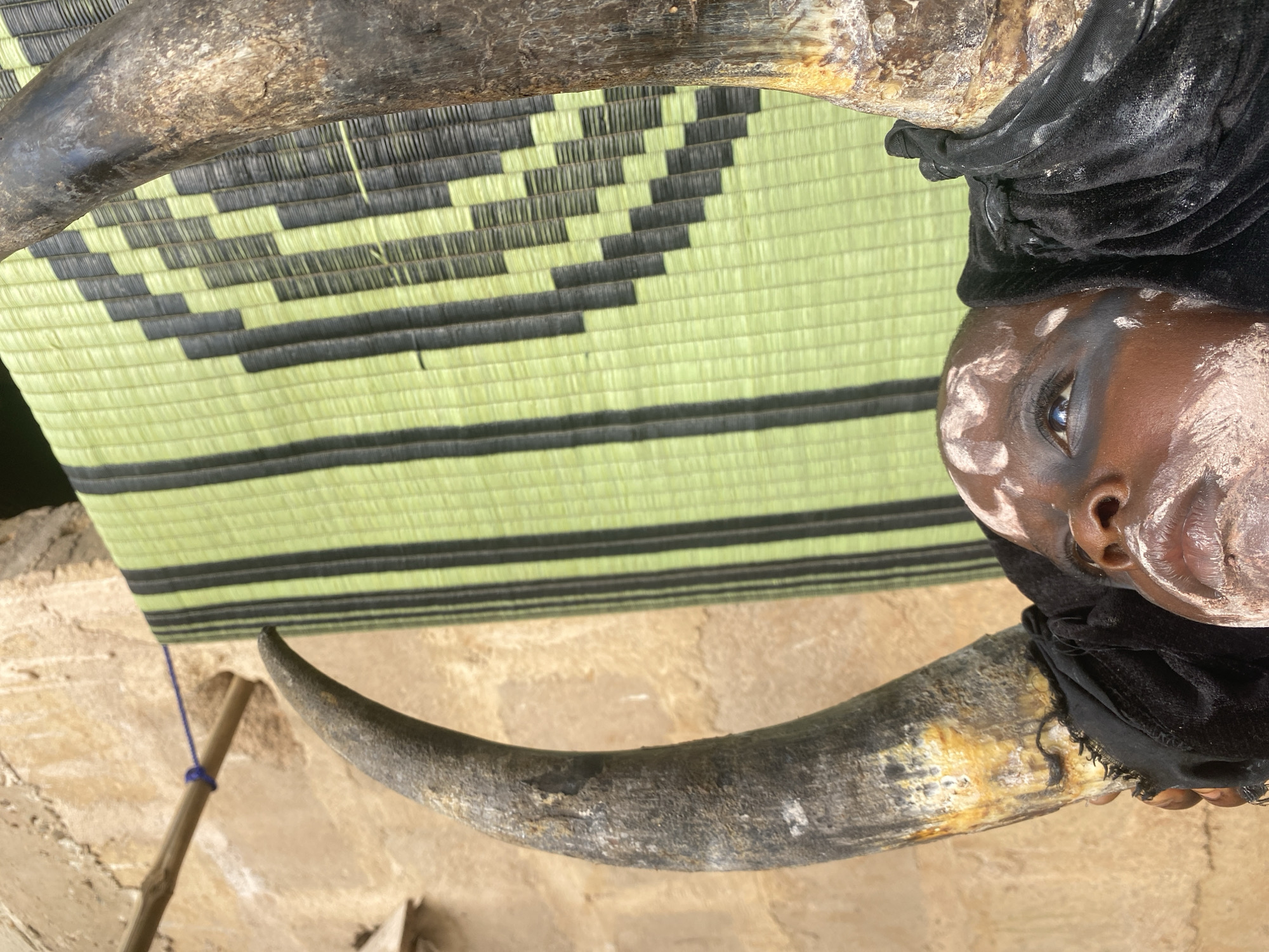 The image depicts a person, possibly a member of the Ekpe society, in Ufu Esioni, Nigeria. The person is adorned with traditional white face paint, wearing a black cloth headdress, and positioned between two large, curved animal horns. The person lies on a mat with alternating green and dark green woven squares. A wooden pole tied with a blue string lies partially visible on a dirt floor behind the person. The scene takes place outdoors, presumably in a rural or village setting.