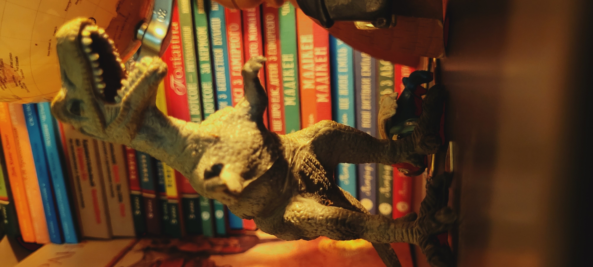 In a dimly lit indoor setting, a plastic toy dinosaur stands on a dark, reflective surface, its head turned to the right. Behind the dinosaur, a stack of colorful books forms a backdrop, their titles mostly obscured. To the right of the dinosaur, a portion of a globe is visible, depicting geographical locations. A few other small toy figures are scattered around the dinosaur's feet. The lighting suggests a low-light environment, with dramatic shadows cast by the dinosaur and the surrounding objects.