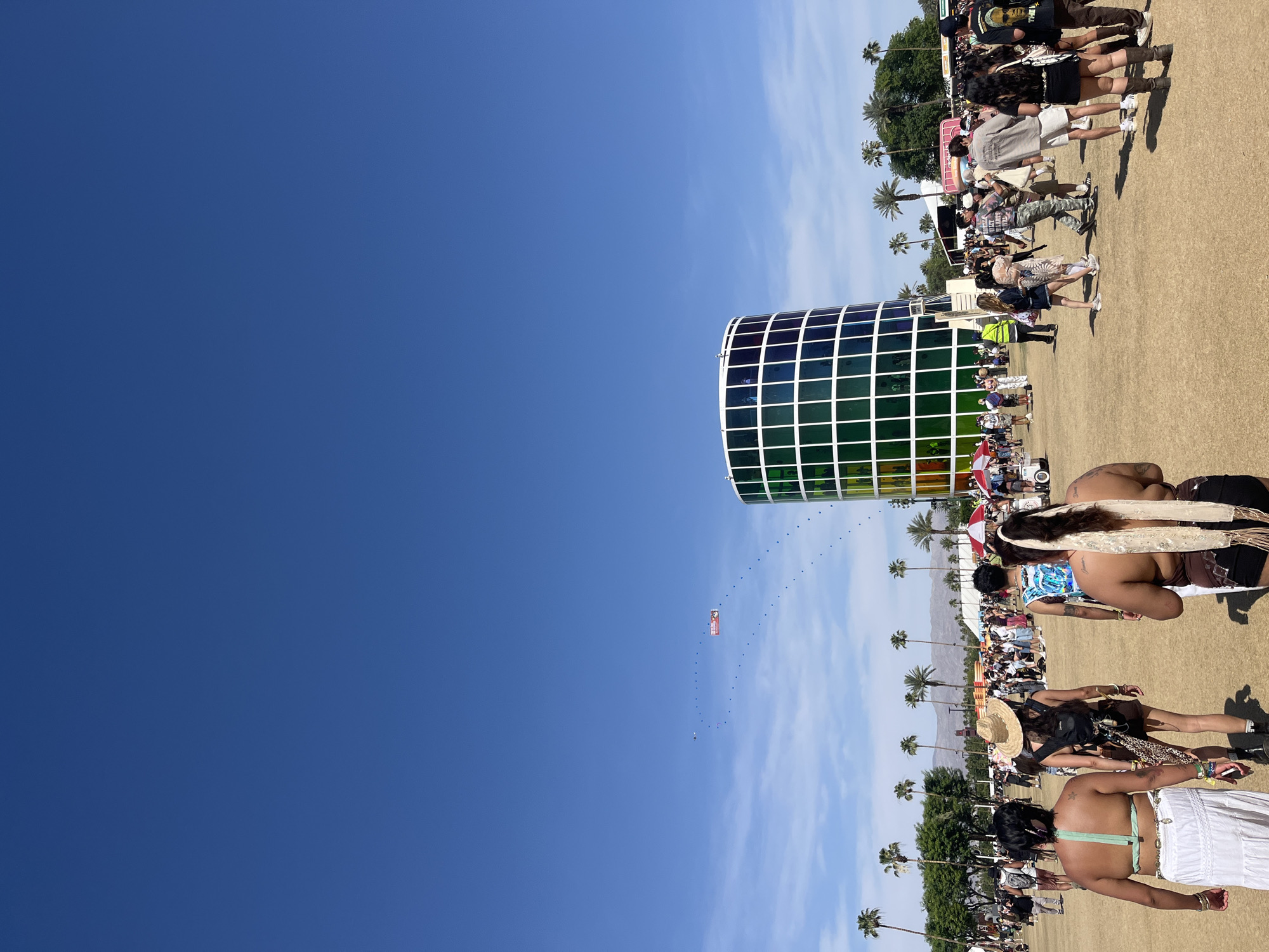 The image depicts a daytime scene in Indio, United States of America. The sky is predominantly blue, taking up approximately half of the image. The right side of the image shows a crowd of people. A large, multi-colored, cylindrical art installation is located in the mid-ground. Several palm trees are interspersed within the crowd and landscape.