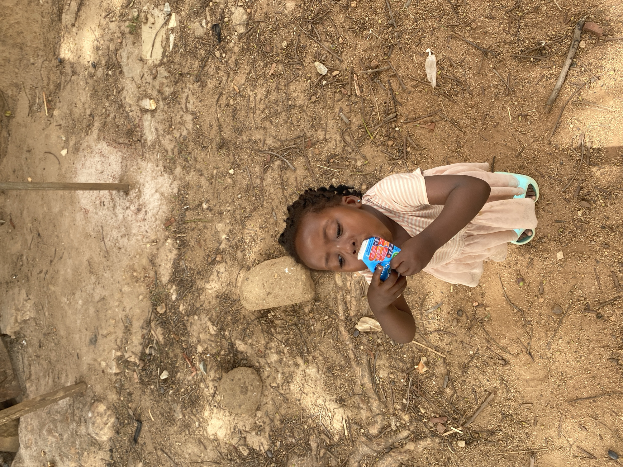 A young Black girl lies on the ground outside, presumably in Kuru Dungus, Nigeria. She is wearing a light pink dress with white stripes and light blue sandals. She is drinking from a blue juice box. The ground is primarily dirt with small sticks and debris scattered around. A couple of stones are near her head. In the upper left, there's a concrete-like patch with a wooden support structure. The lighting indicates daytime.