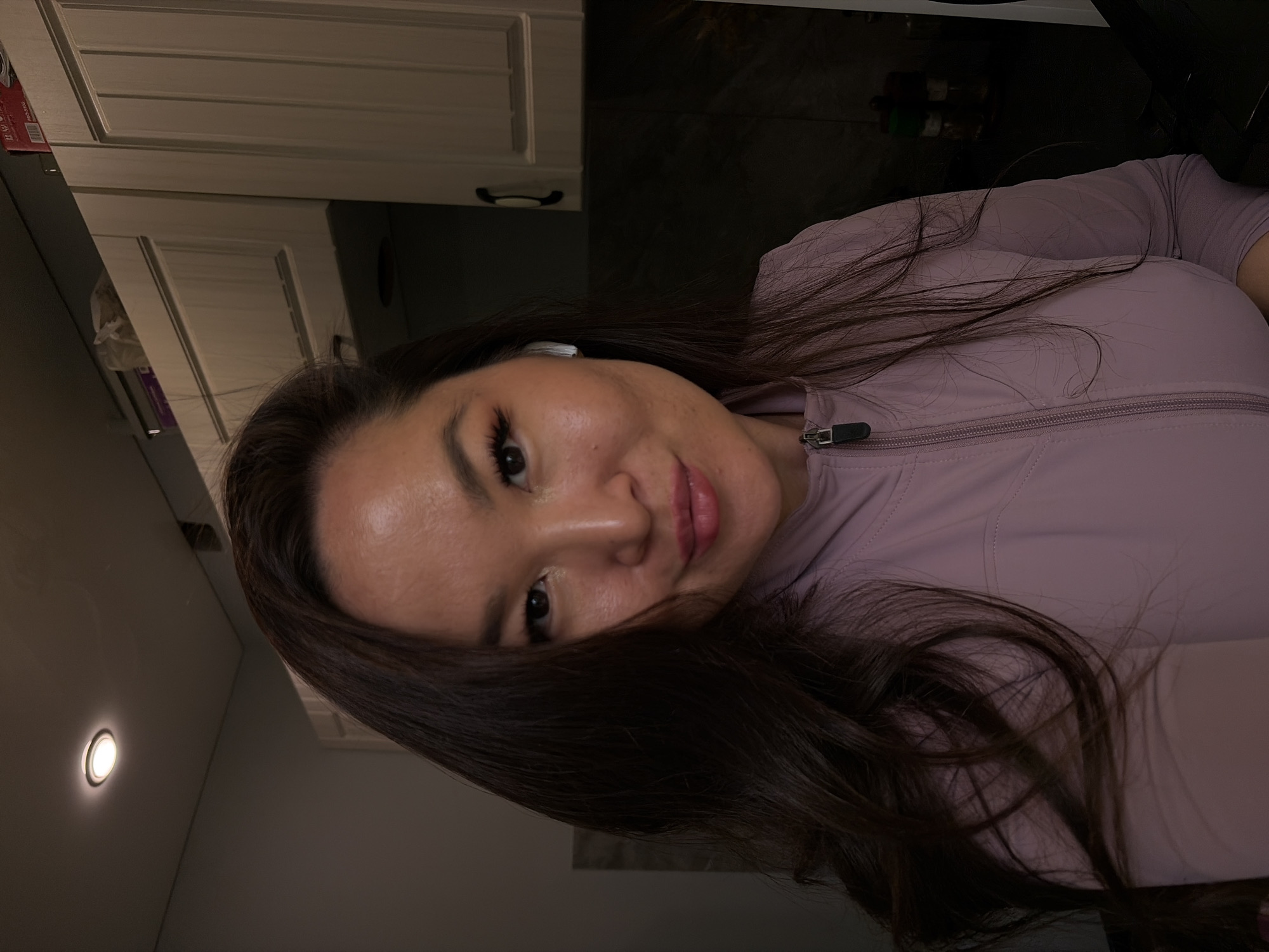 The media file shows a close-up, rotated selfie of a woman with long, dark brown hair. She is wearing a light purple zip-up jacket. Her makeup includes dark eyeliner and mascara, and her lips are a soft pink. She appears to be indoors, with kitchen cabinets visible in the background. A circular light fixture is also visible on a wall. The lighting suggests it could be evening or a dimly lit indoor space. The woman is looking at the camera with a slight smile, conveying a casual and relaxed emotion. There is no visible text in the image. The context provided indicates the location is Chaikovsky, Russia.