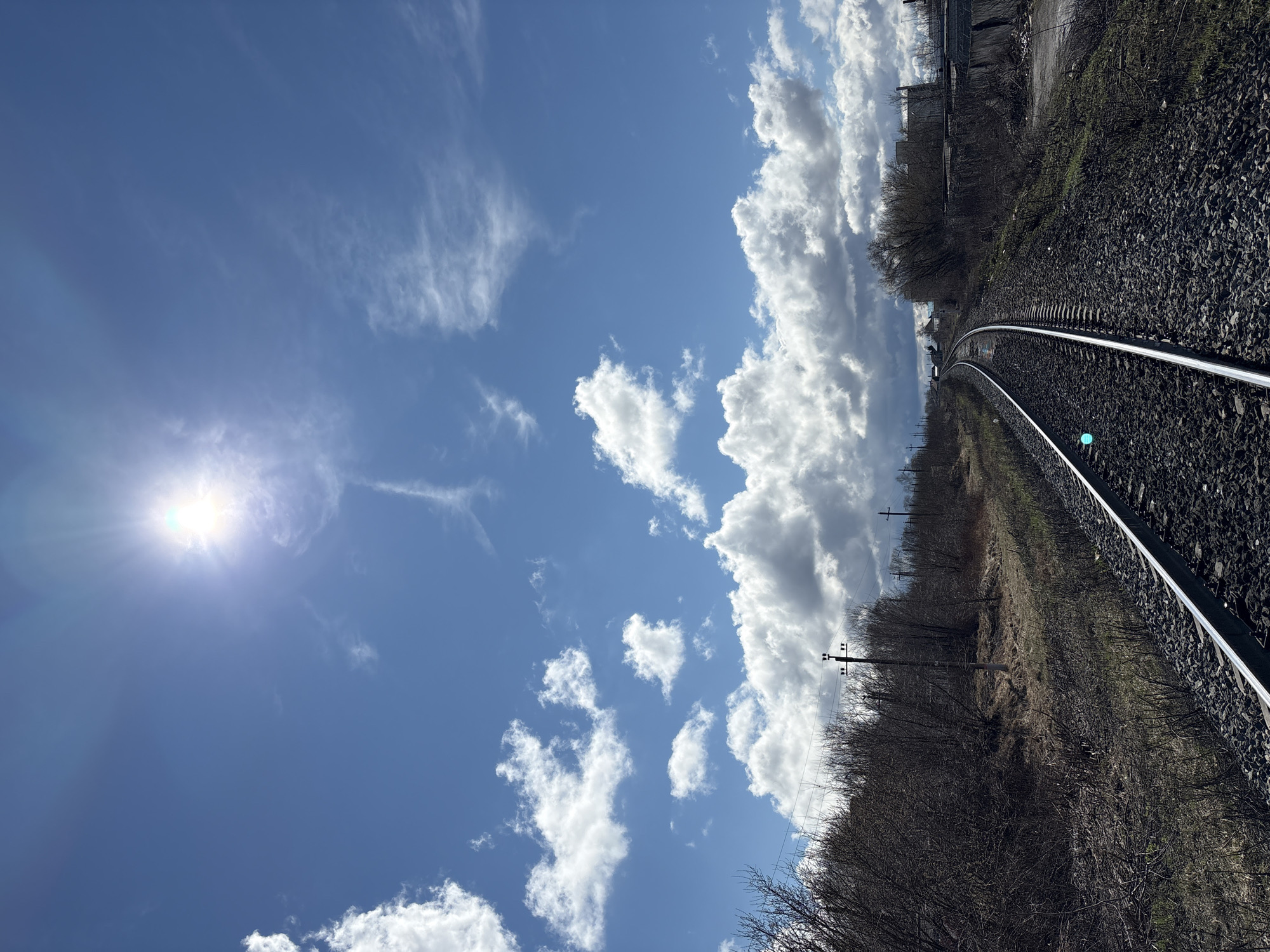 The image depicts a railway track curving towards the horizon under a blue sky with scattered clouds. The sun is visible in the sky, emitting a bright light. The railway track is bordered by gravel and vegetation, with bare trees and some buildings visible in the distance. The location is Ulyanovsk, Russia. There are no people visible in the frame.