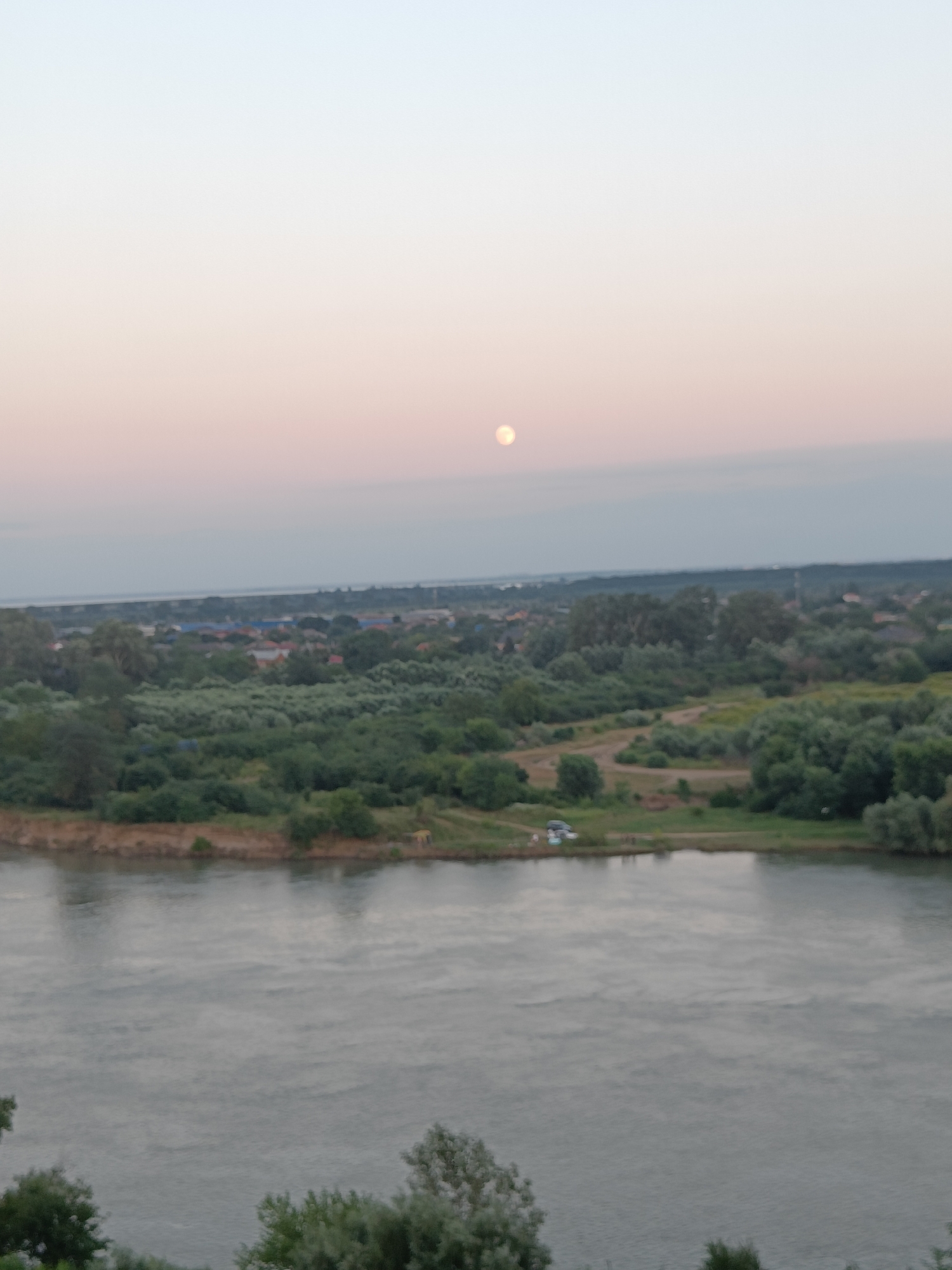 The image depicts a serene landscape scene in Krasnodar, Russia, likely taken during the twilight hours. A vast river dominates the foreground, its surface reflecting the soft, pastel hues of the sky. The sky itself transitions from a pale blue at the horizon to a soft pink and lavender near the top of the frame, suggesting either sunrise or sunset. A faint, luminous disc, possibly the moon or a distant sun, is visible high in the sky.
The middle ground is characterized by a lush, green expanse of trees and vegetation. Beyond the dense greenery, a scattered settlement with visible rooftops and structures is nestled. The overall impression is one of tranquility and rural charm.
On the riverbank in the middle distance, a few small vehicles are parked, and what appear to be a few individuals are gathered, though they are not distinct enough to discern any specific activity. The presence of these elements suggests human presence and perhaps a leisurely outing by the river. The weather appears calm, with no indication of strong winds or precipitation. The setting evokes a sense of peace and the beauty of nature at the edge of a settlement. No visible text is present in the image.