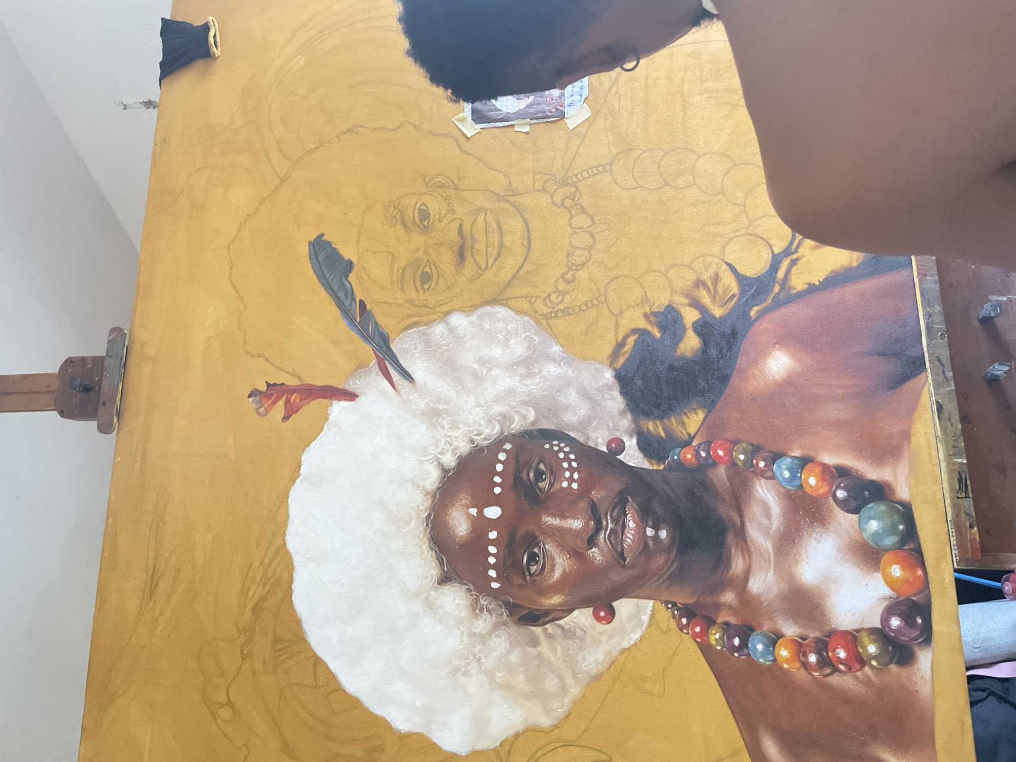 The image shows an artist actively working on a large-scale painting, likely in an art studio in Ado, Nigeria. The artist's back of the head, with dark curly hair, and their right arm are visible from a top-down perspective, indicating they are positioned over the canvas.
The painting itself features a highly detailed portrait of a dark-skinned individual with white facial markings, including dots and lines across the forehead, cheeks, and chin. This figure is adorned with a voluminous, white, textured headdress or hairpiece, from which a red-tipped feather emerges. A prominent, multi-colored beaded necklace, comprising large, spherical beads in various shades of red, orange, green, blue, brown, and purple, encircles the person's neck. The painted figure's expression is calm and direct.
In the background of the canvas, a larger, secondary figure is outlined in pencil sketch, also depicting a smiling person with a suggested beaded necklace. The canvas itself has a rich, golden-yellow base color. A small reference image, partially taped to the canvas near the top, is visible with the text "The Young Man". An easel supports the canvas on the left, and faint pencil lines indicating further design elements are visible on the unpainted areas. The image captures a moment of creative process, with one figure largely completed in detail and another still in its preliminary sketch phase.